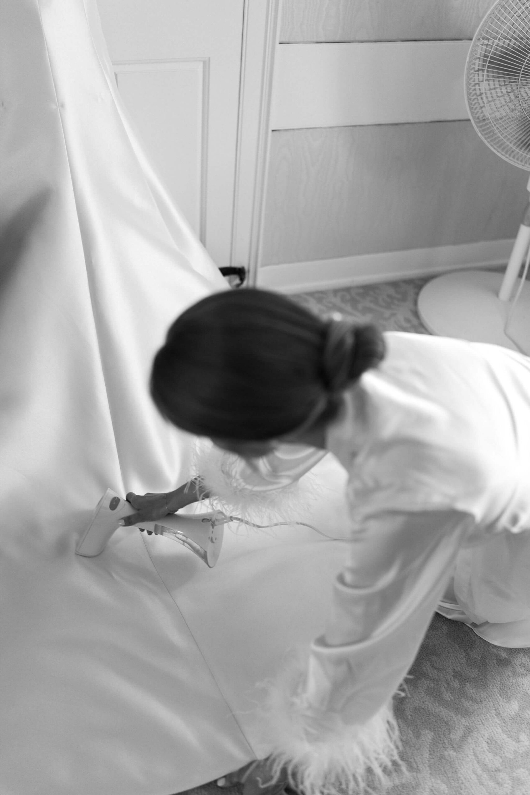 black and white image of a bride (in a robe with fuzzy cuffs) kneeling to steam the train of her wedding dress