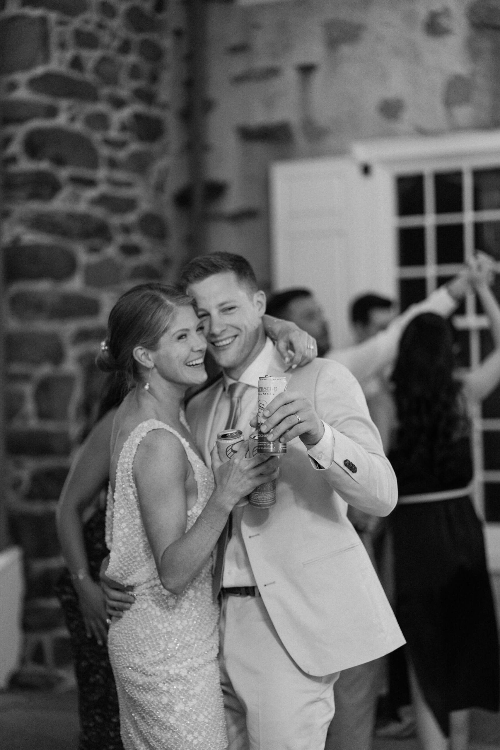 black and white image of bride and groom embracing and smiling during their wedding reception, both holding drinks