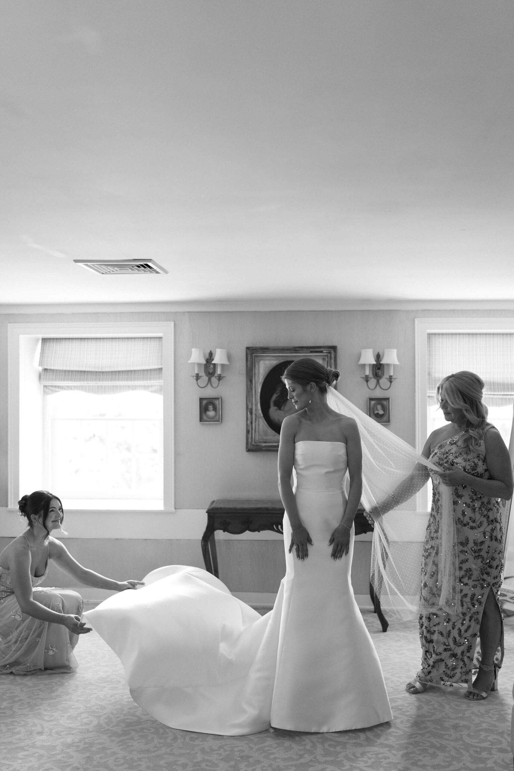 black and white image of a bride in her wedding gown looking over her shoulder while her sister (left) kneels to straighten her train, and her mother (right) holds her veil