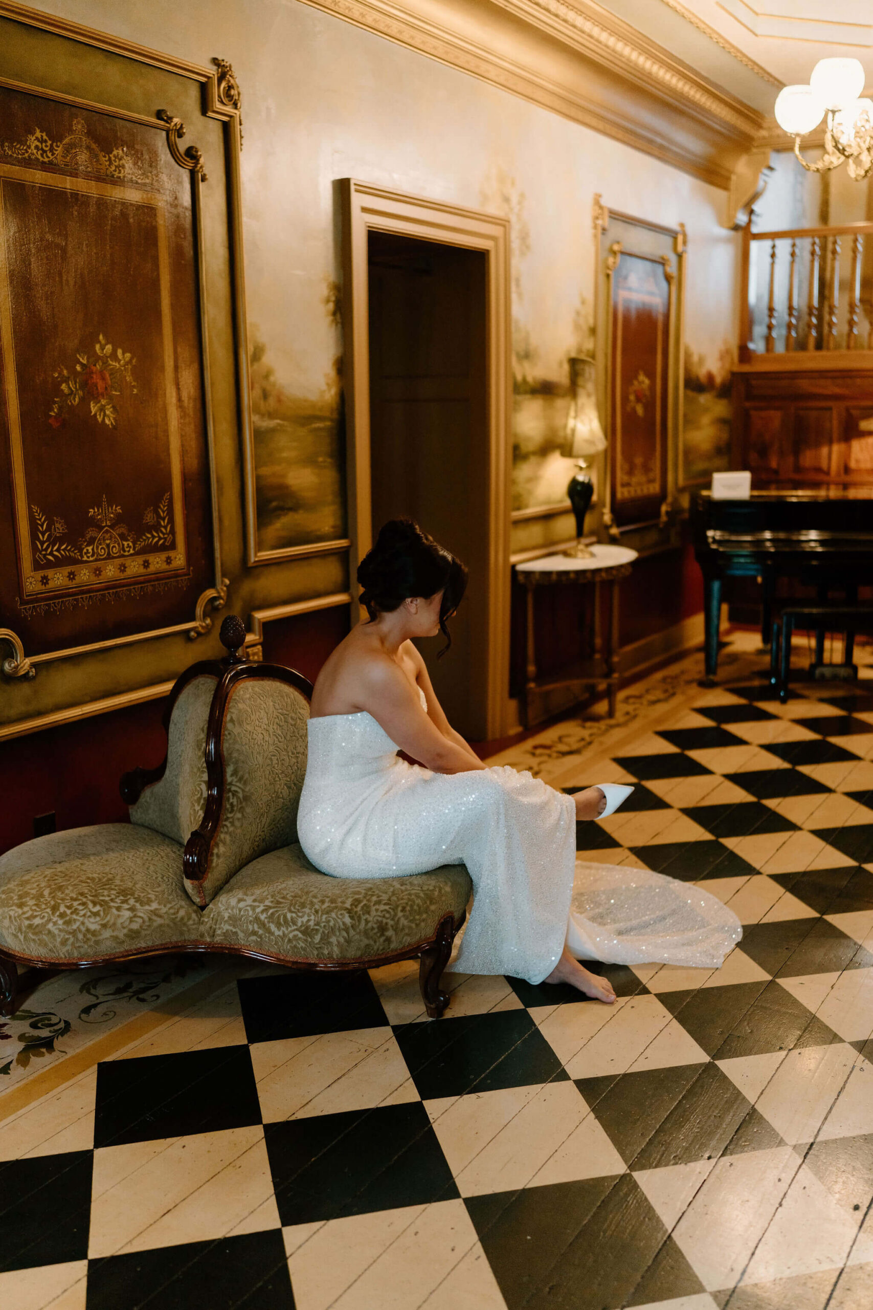 bride seated in an ornate entryway with black and white checkered flooring, putting on her heels