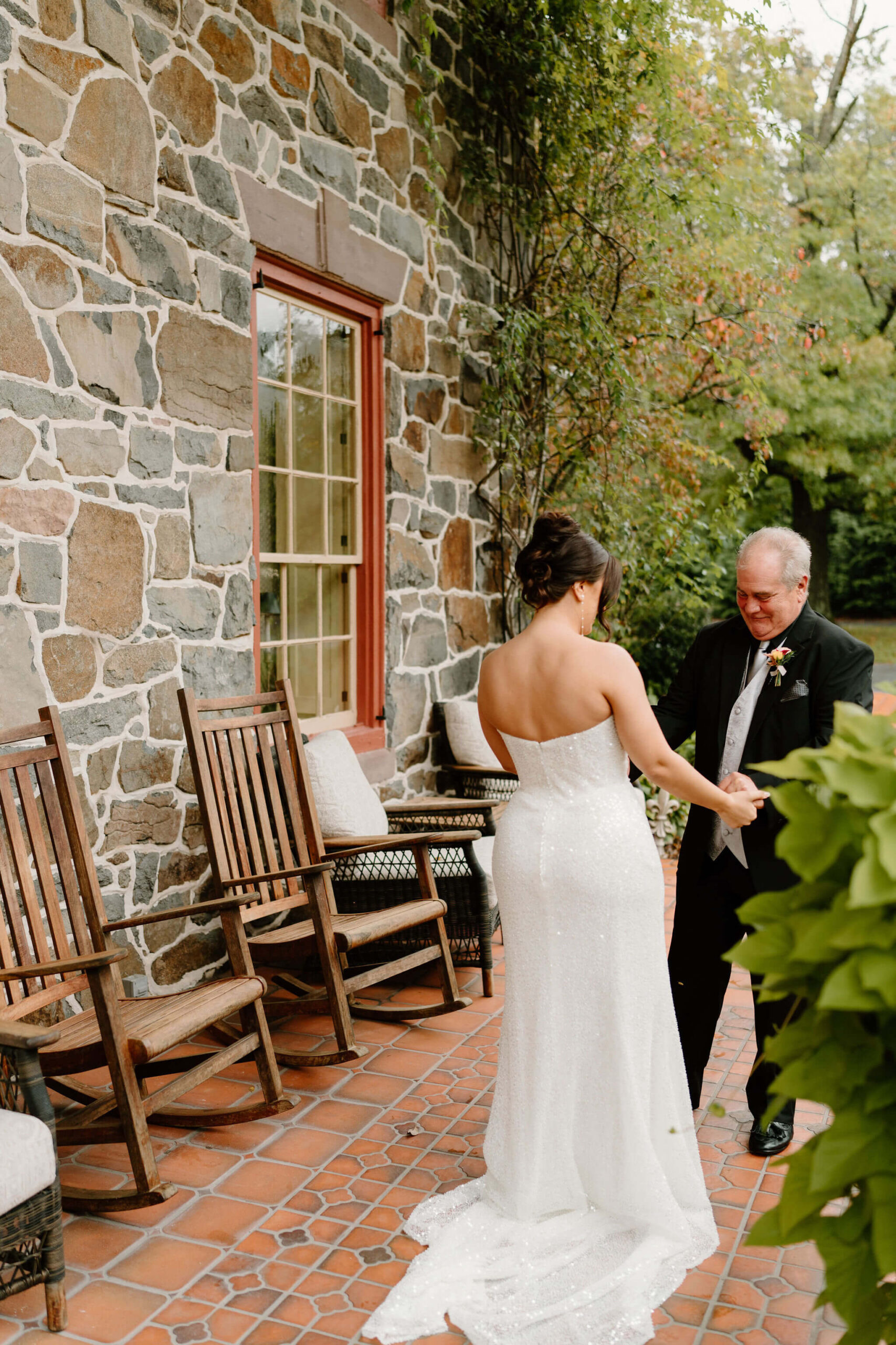 father of the bride admiring his daughter's sparkling wedding gown after their first look on her wedding day, against a stone home background
