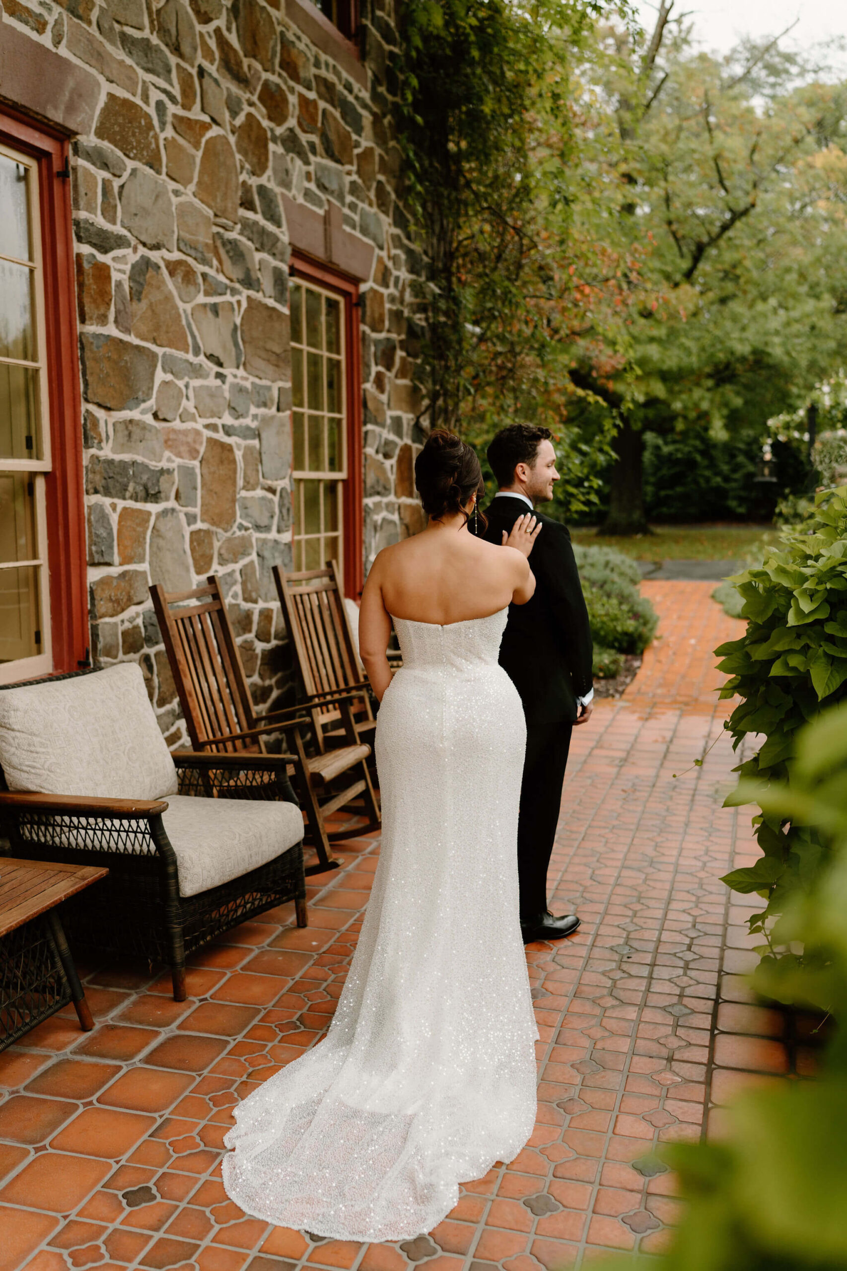 bride in a sequined wedding dress tapping her groom (black suit) on the shoulder during their first look on a tiled porch with a stone background