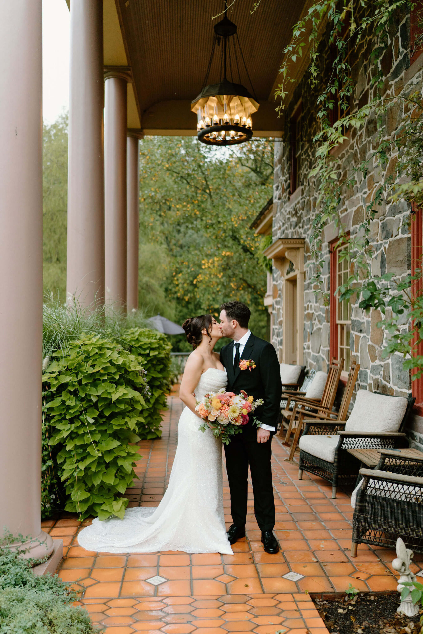 bride and groom kissing on a covered tile porch against a stone wall with potted greenery, while she holds her colorful bridal bouquet