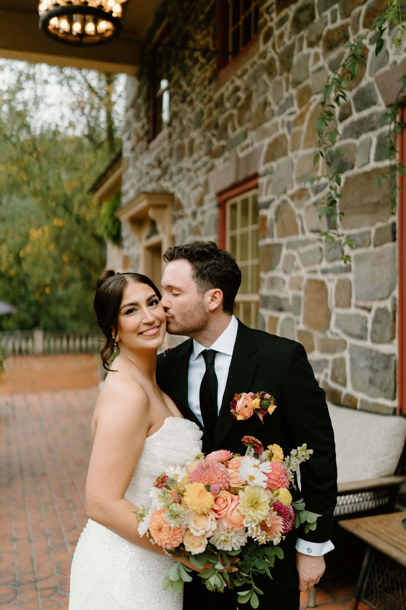 groom kissing bride on the cheek as she smiles at the camera while holding her colorful bridal bouquet in front of a historic estate