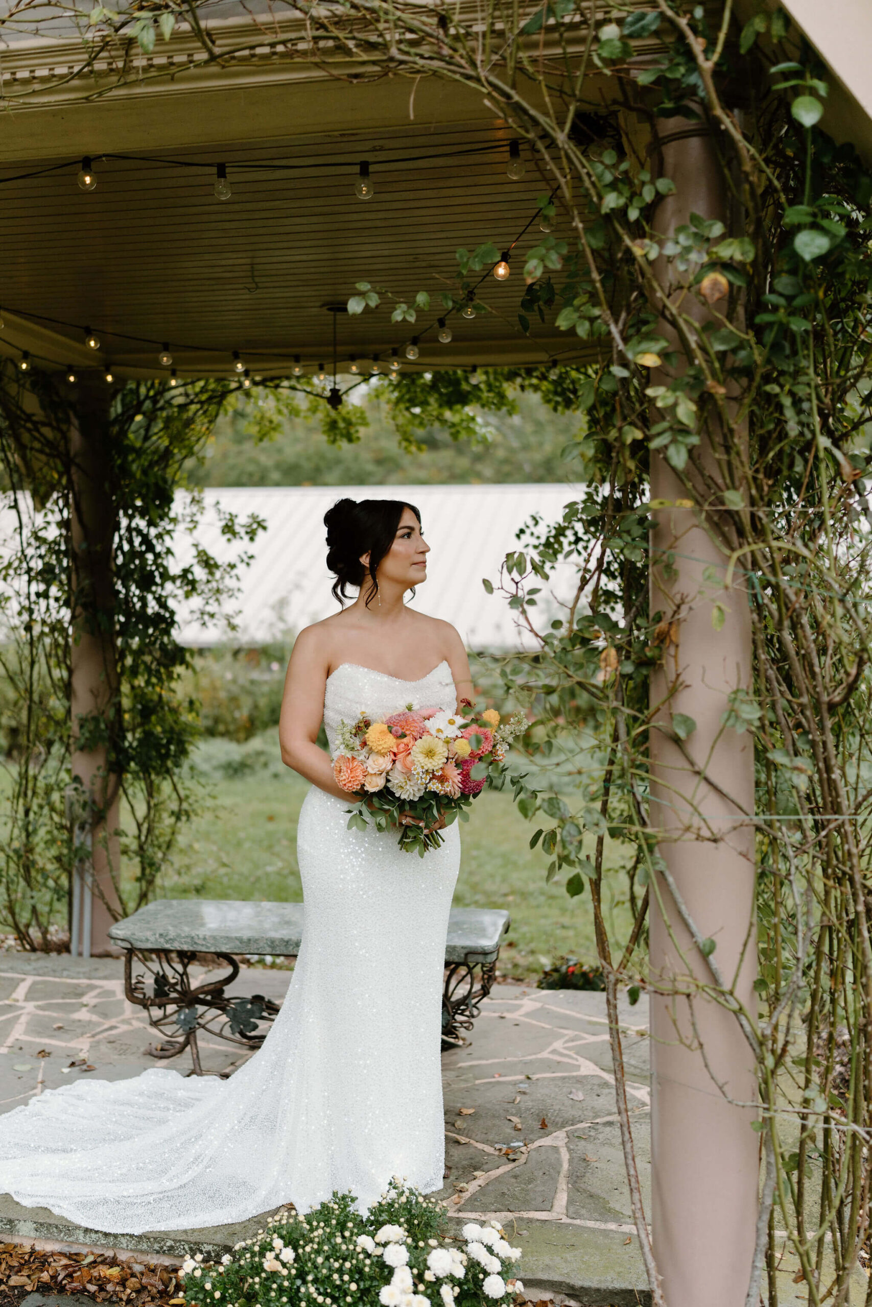 bride in a sparkly, sequined wedding gown holding her colorful bridal florals and looking off camera, standing beneath a covered gazebo with crawling vines