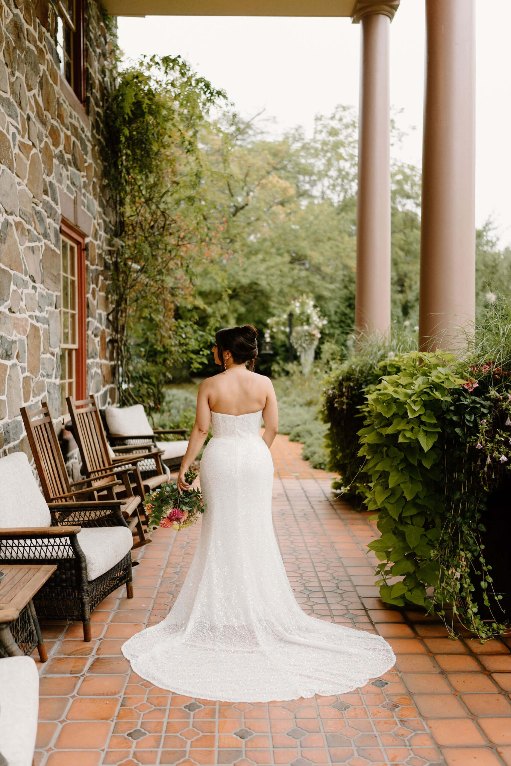 portrait of the back of the bride's sparkly, sequined wedding gown as she stands on a tiled covered porch against a stone wall, her colorful bouquet held at her side