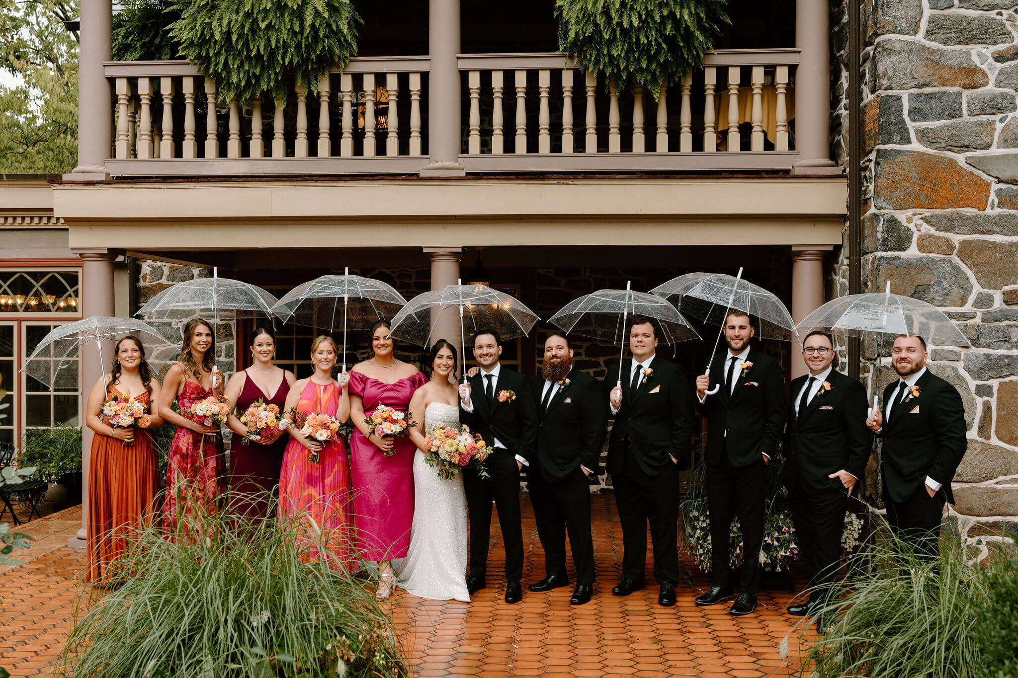 bride, groom, and wedding party gathered on a tile porch beneath clear umbrellas, smiling at the camera