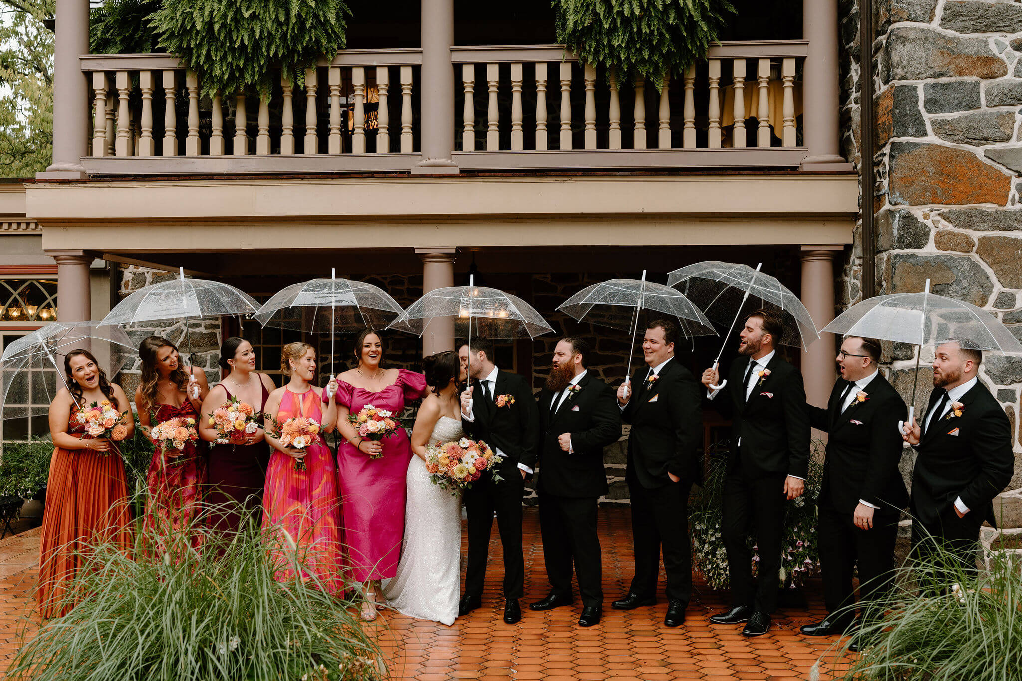 bride, groom, and wedding party gathered on a tile porch beneath clear umbrellas, smiling at the bride and groom as they kiss