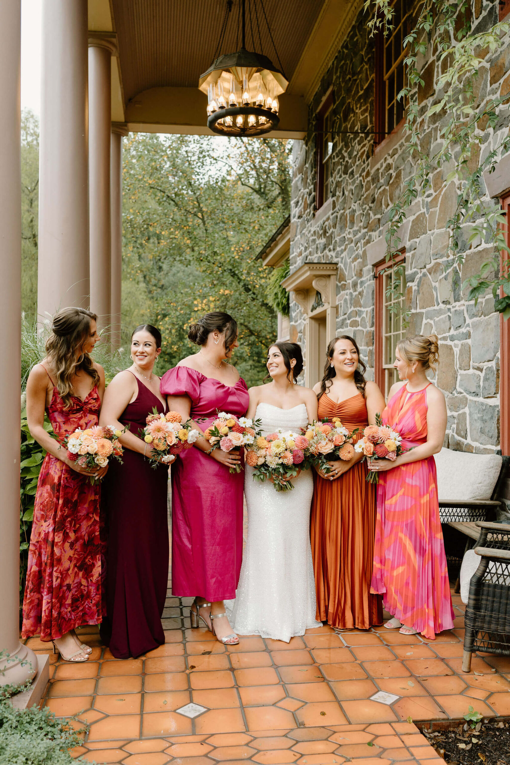 bridesmaids with bride in coordinated, mismatched wedding dresses of oranges, pinks, and reds, holding their flowers and looking at each other, smiling