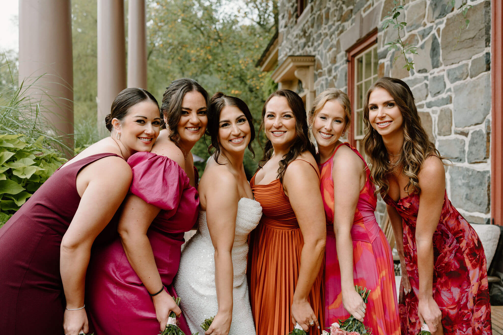 bridesmaids with bride in coordinated, mismatched wedding dresses of oranges, pinks, and reds, leaning together and smiling at the camera
