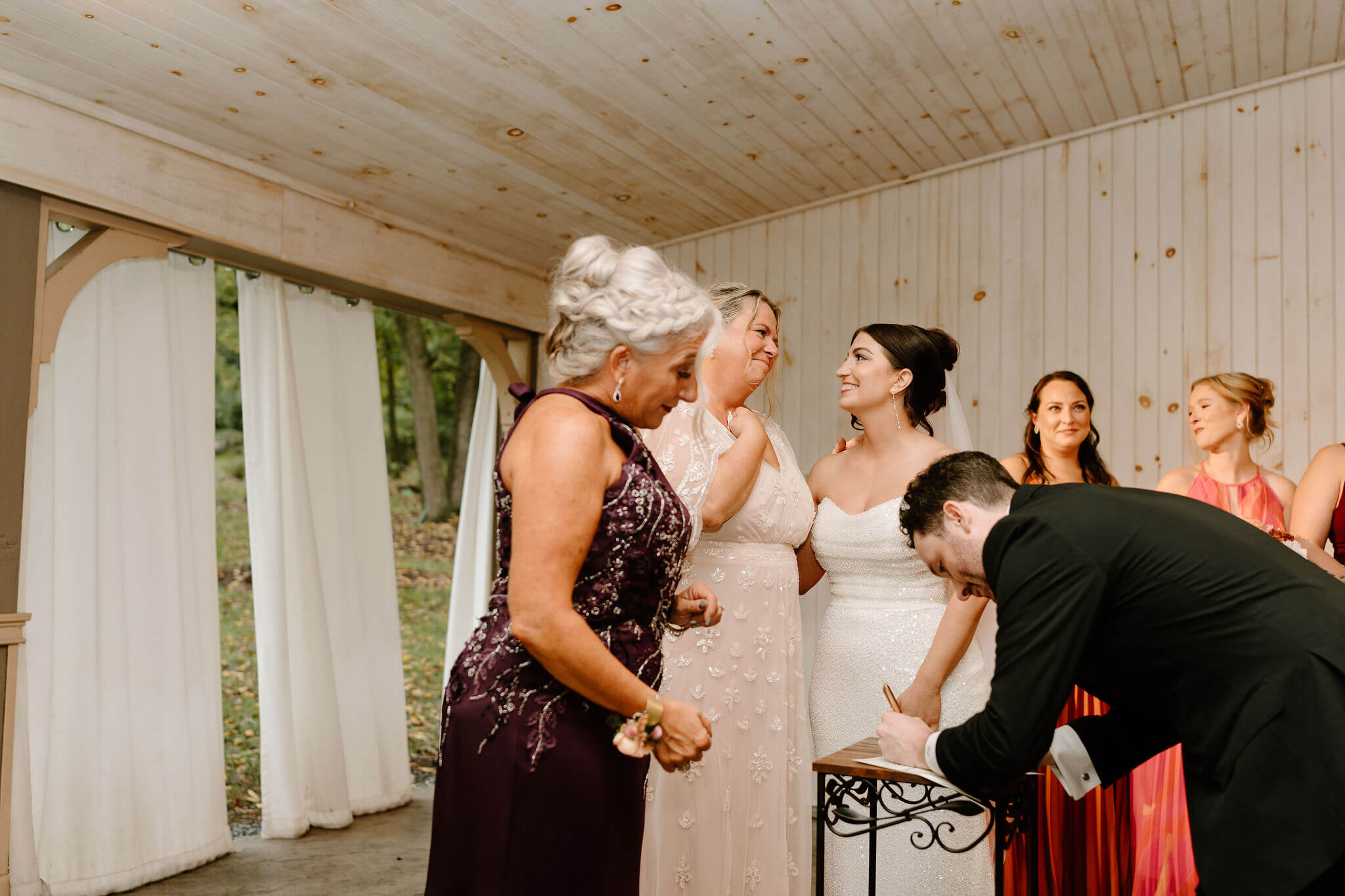 bride and her mother embracing with her mother's hand over her heart, as the groom and his mother sign the marriage license
