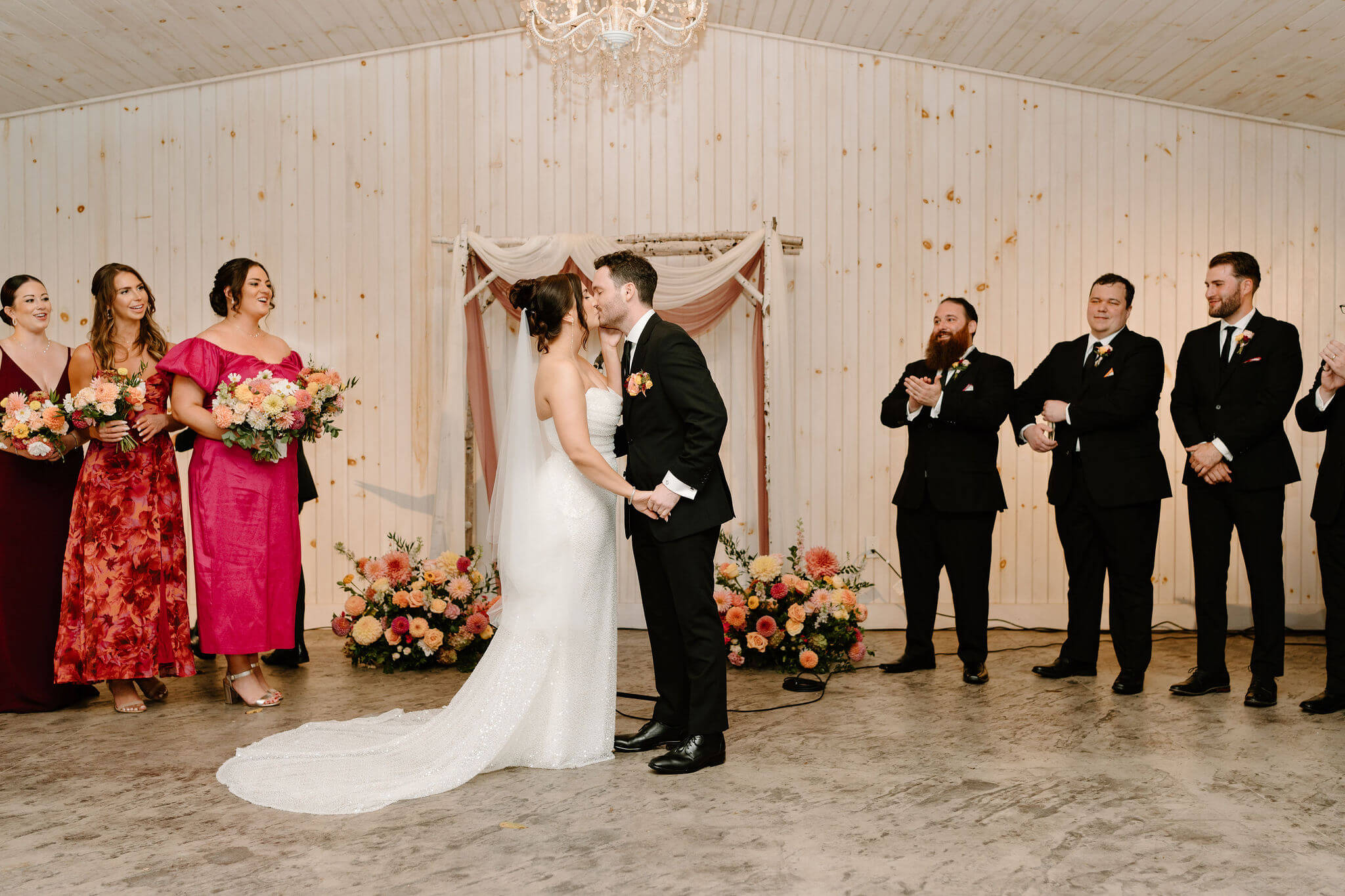 first kiss of bride and groom at the end of their wedding ceremony, wedding party clapping behind them