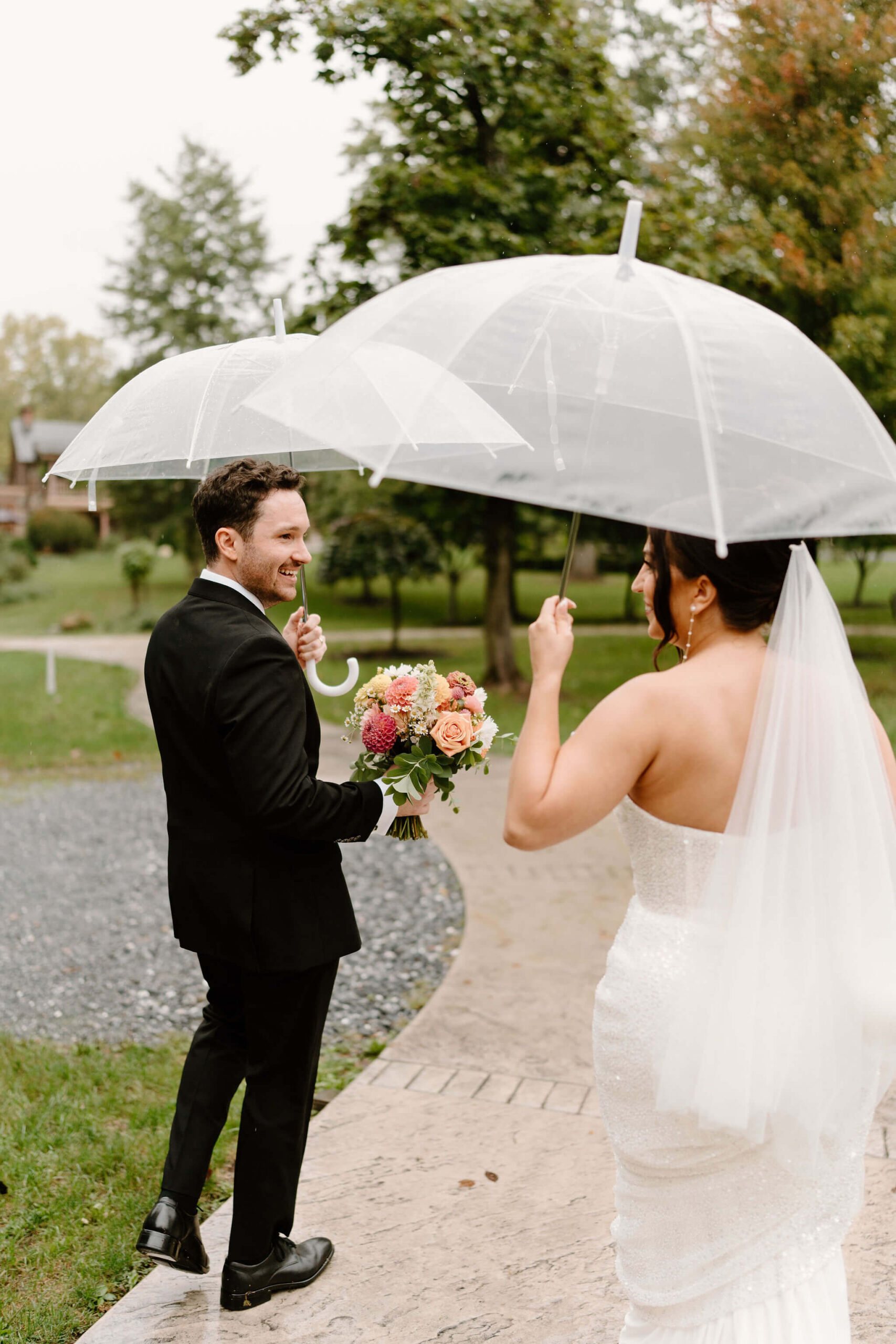 bride and groom exiting ceremony space under clear umbrellas, groom smiling at bride as he holds her bouquet