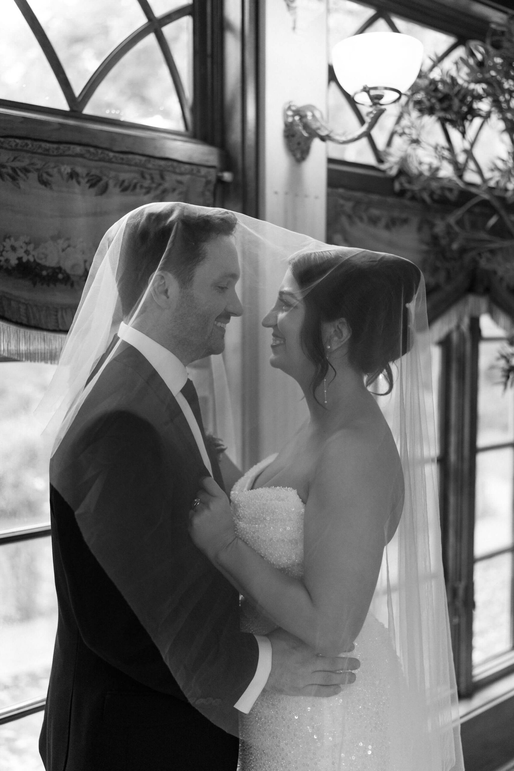 black and white image of bride and groom beneath a wedding veil, smiling at each other