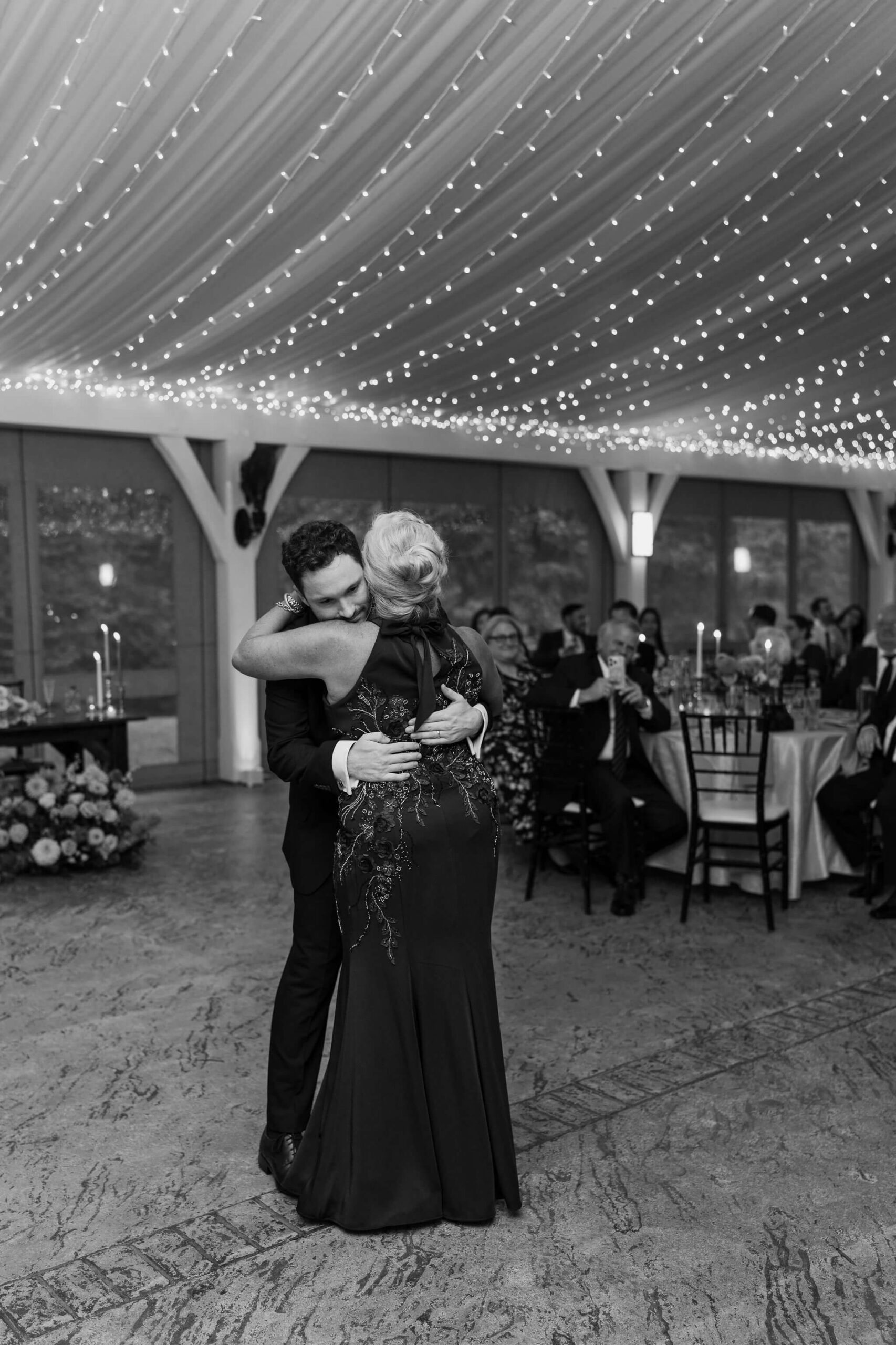 black and white image of a groom and his mother embracing during the mother/son dance at his wedding reception, in a permanent tent structure with lots of string lights