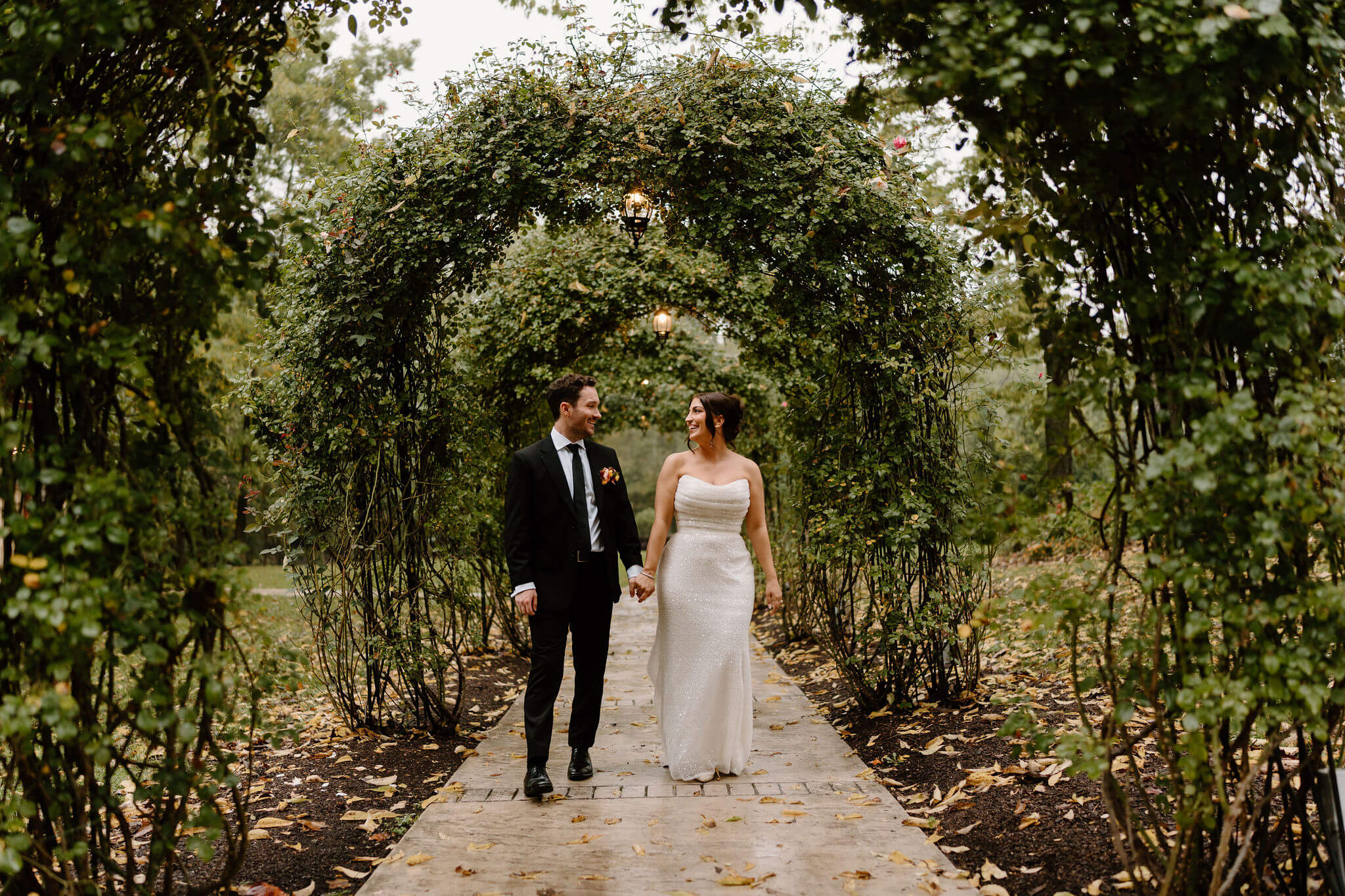 bride and groom holding hands and smiling at each other beneath greenery arbors covering a garden walkway