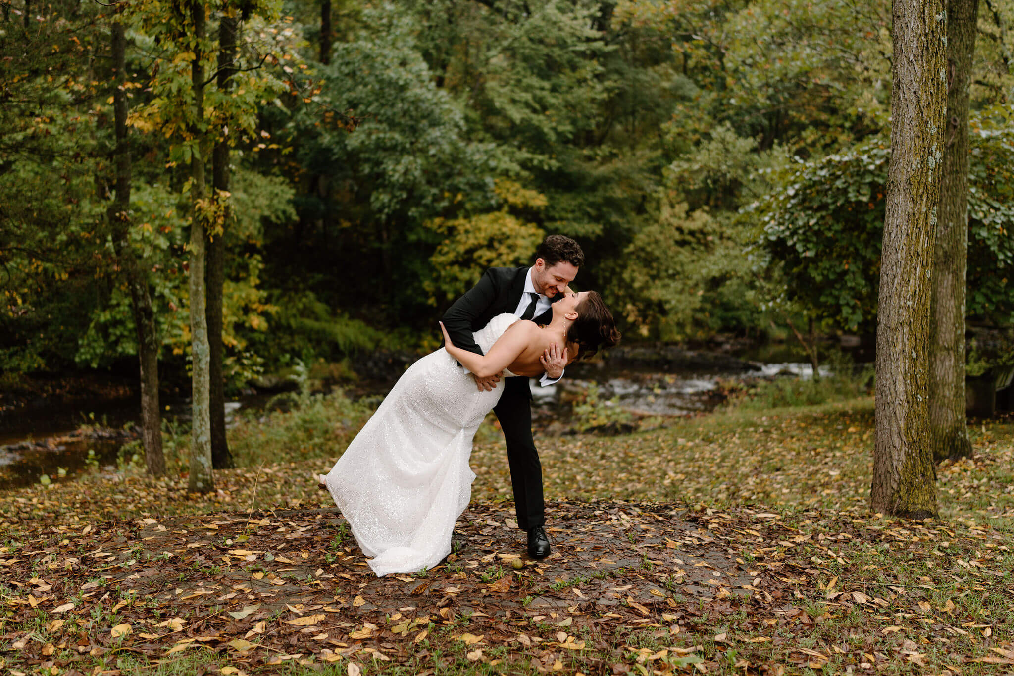 groom dipping bride in a forest setting, a creek in the background and fallen leaves on the ground