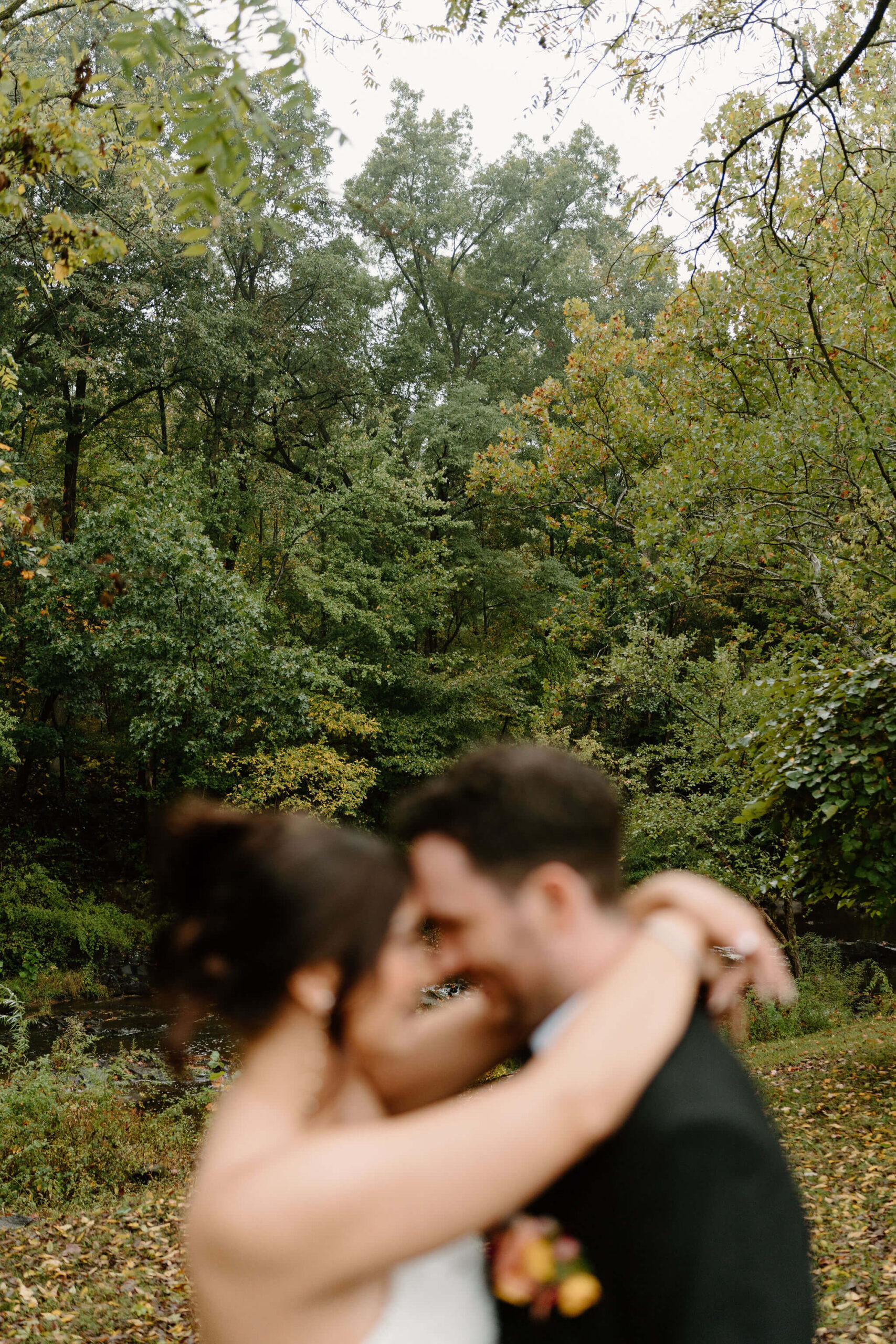 image of bride and groom with their foreheads pressed together, out of focus, while the camera focuses on lush green and fall-colored foliage in the background