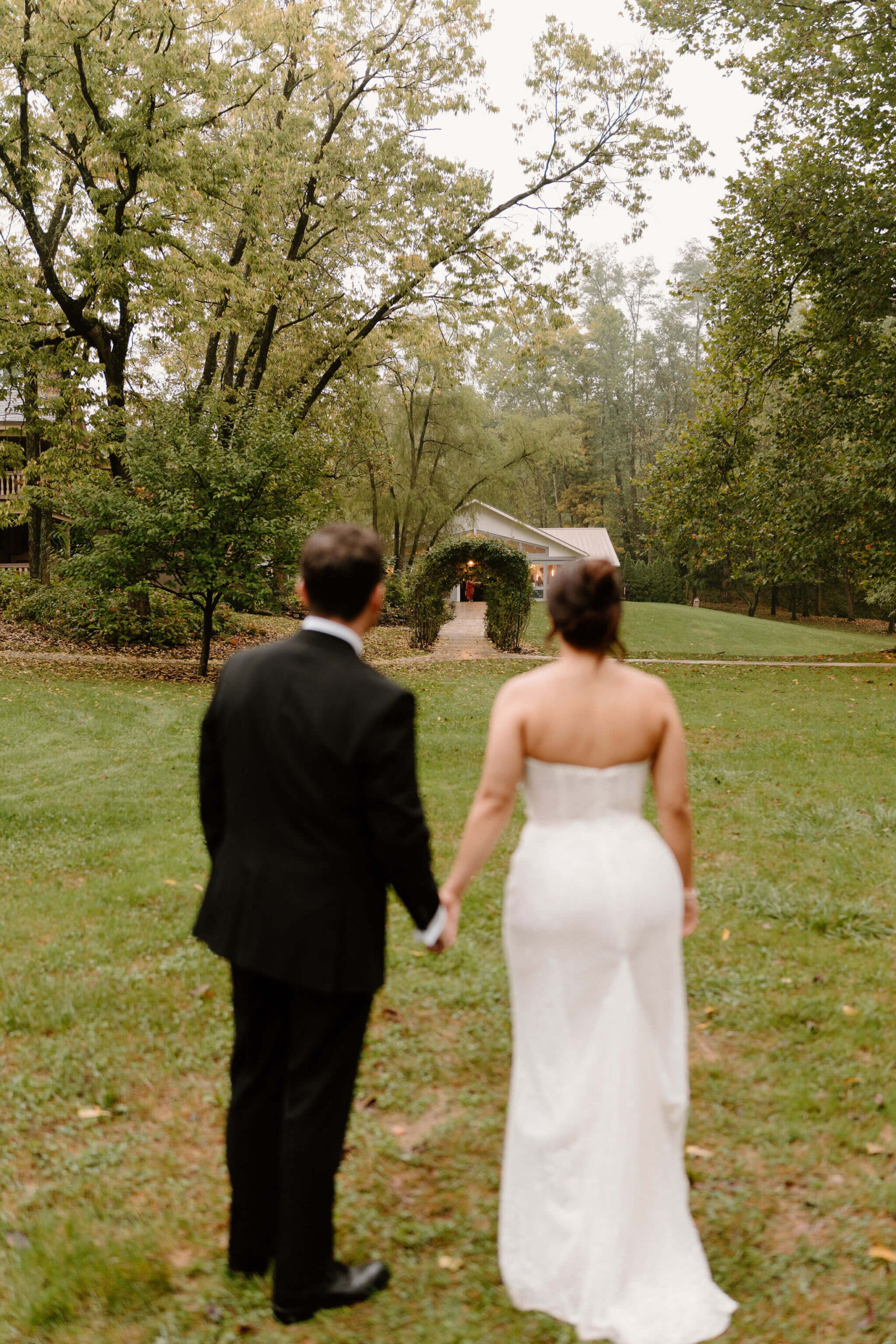bride and groom holding hands (out of focus) looking across a grassy yard and garden path toward their wedding reception venue (in focus)