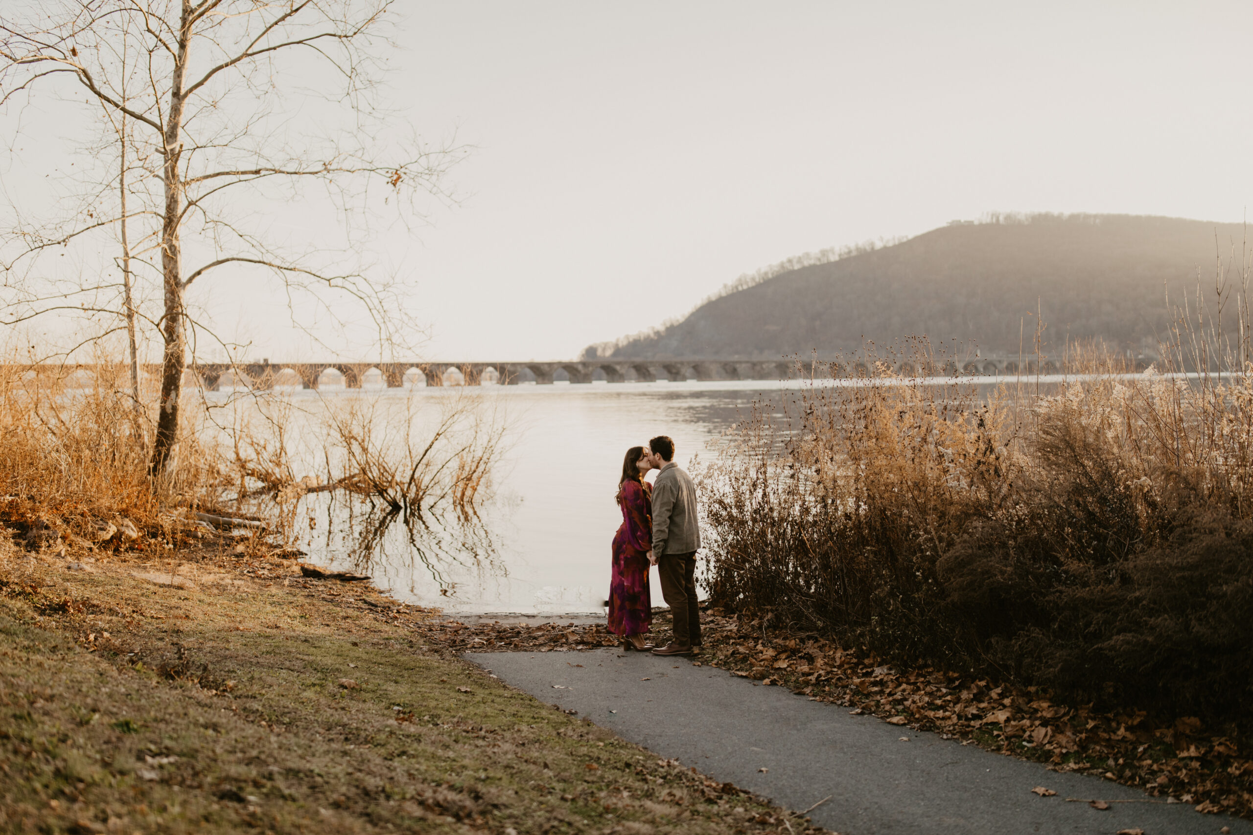 engaged couple kissing, against a winter landscape of a wide river, a mountain in the background and a long bridge spanning the length of the river
