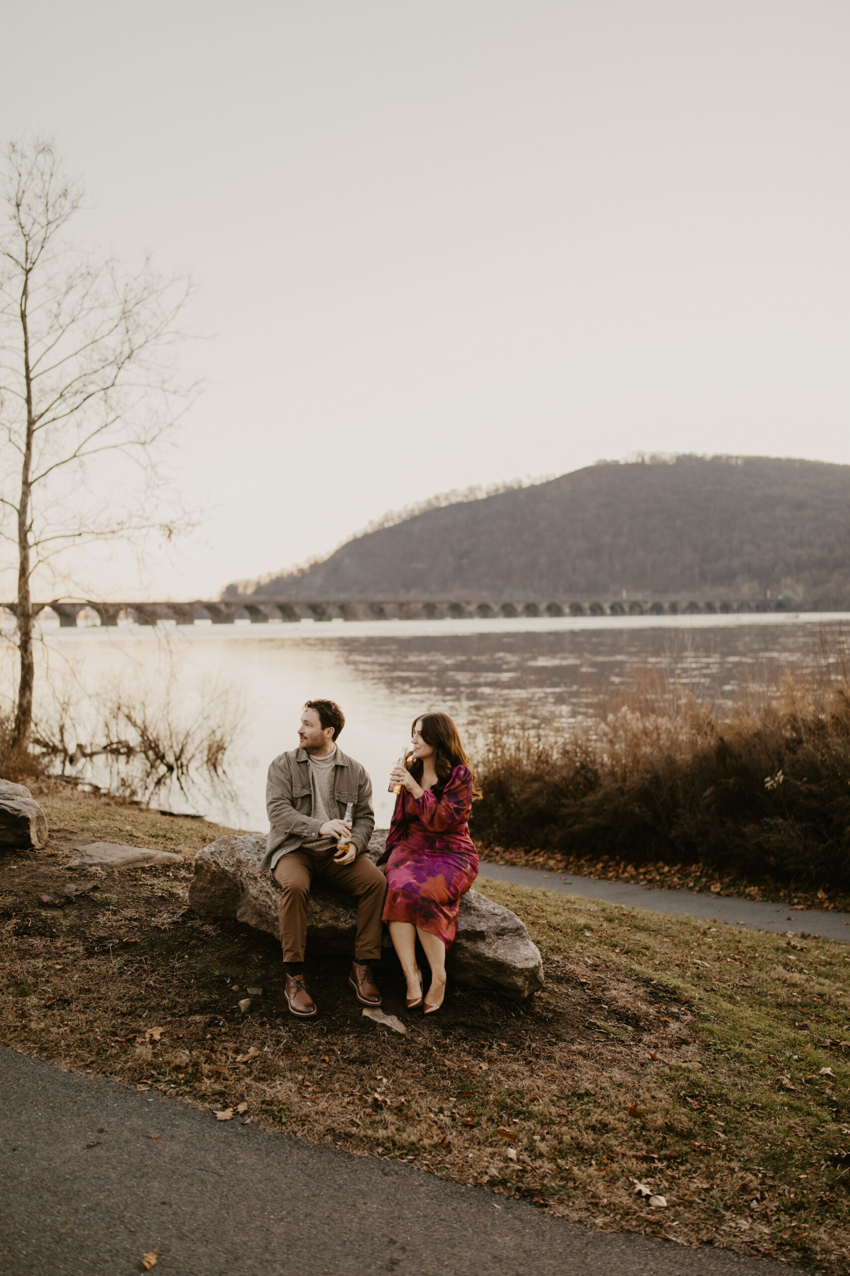 engaged couple sitting on a rock and enjoying a beer, against a winter landscape of a wide river, a mountain in the background
