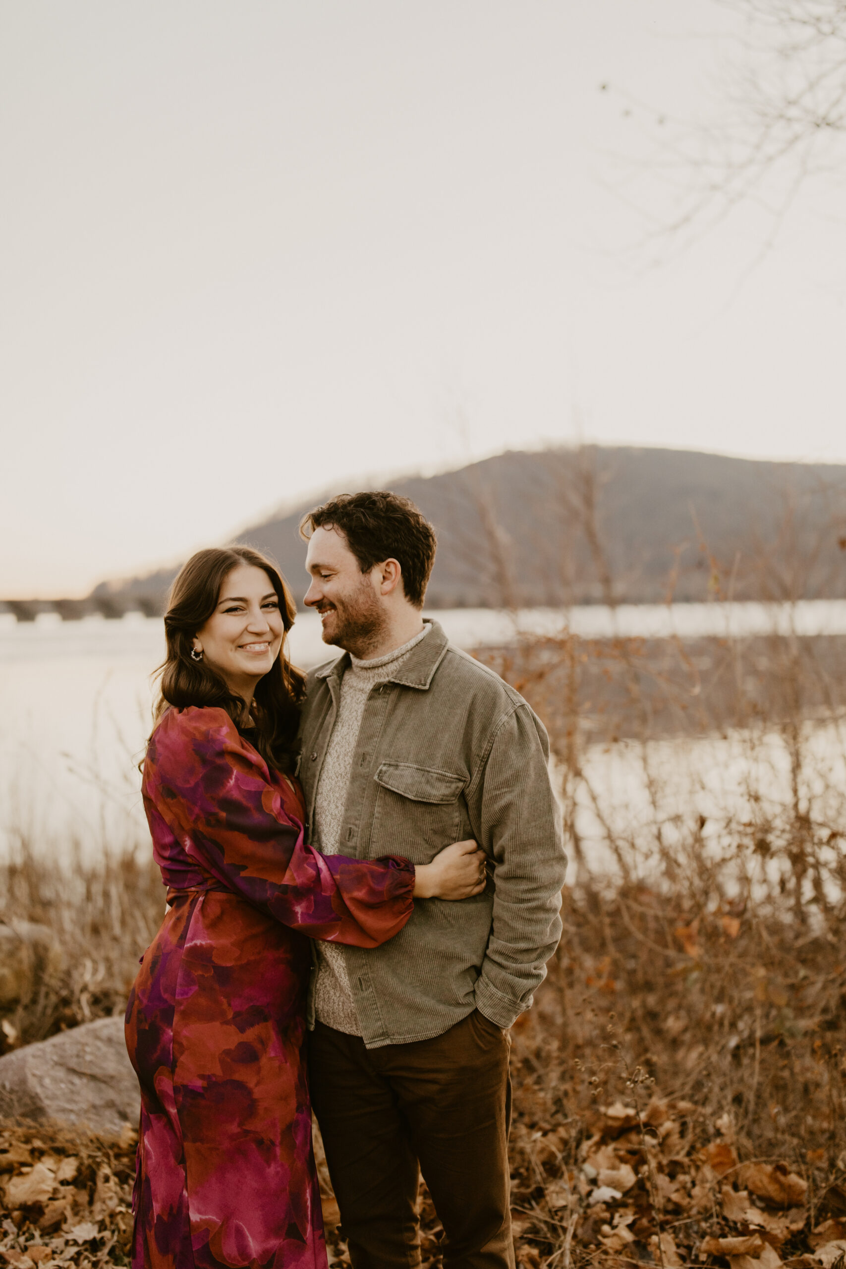 man (brown hair, brown pants, corduroy jacket) smiling at his fiance (long brown hair, colorful red and purple dress) against a winter landscape of a wide river, a mountain in the background