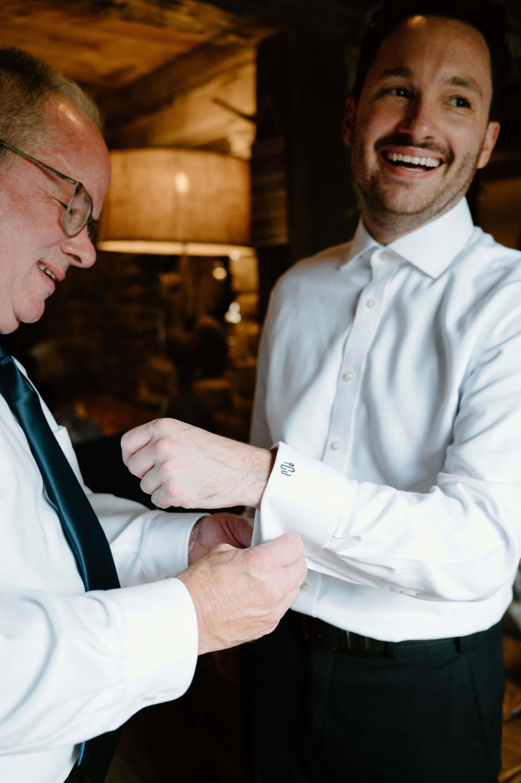father of the groom helping to secure cuff links in the groom's dress shirt, both smiling