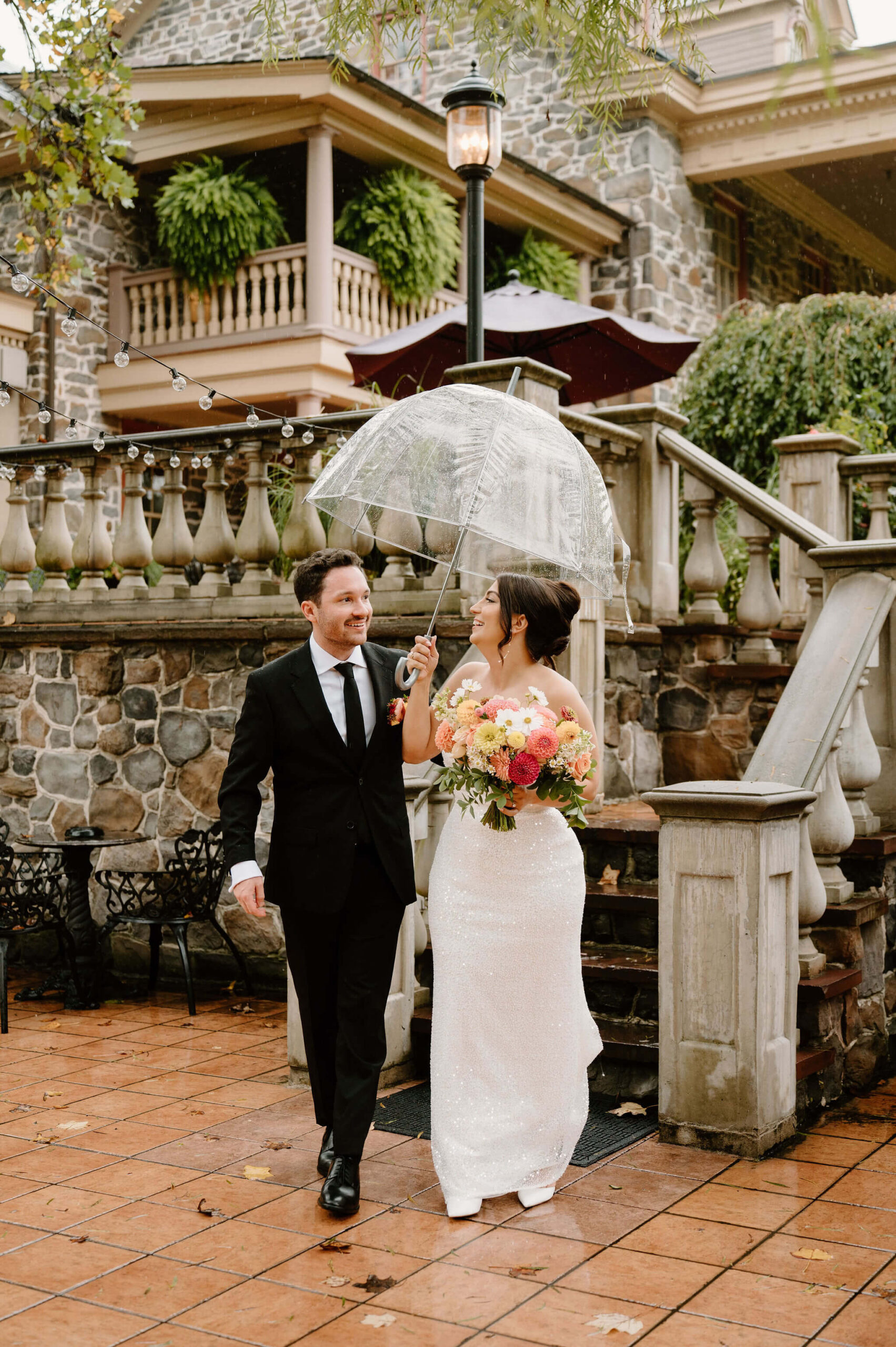 bride and groom walking together down an outdoor stone staircase, away from a large stone mansion, holding an umbrella and smiling toward each other