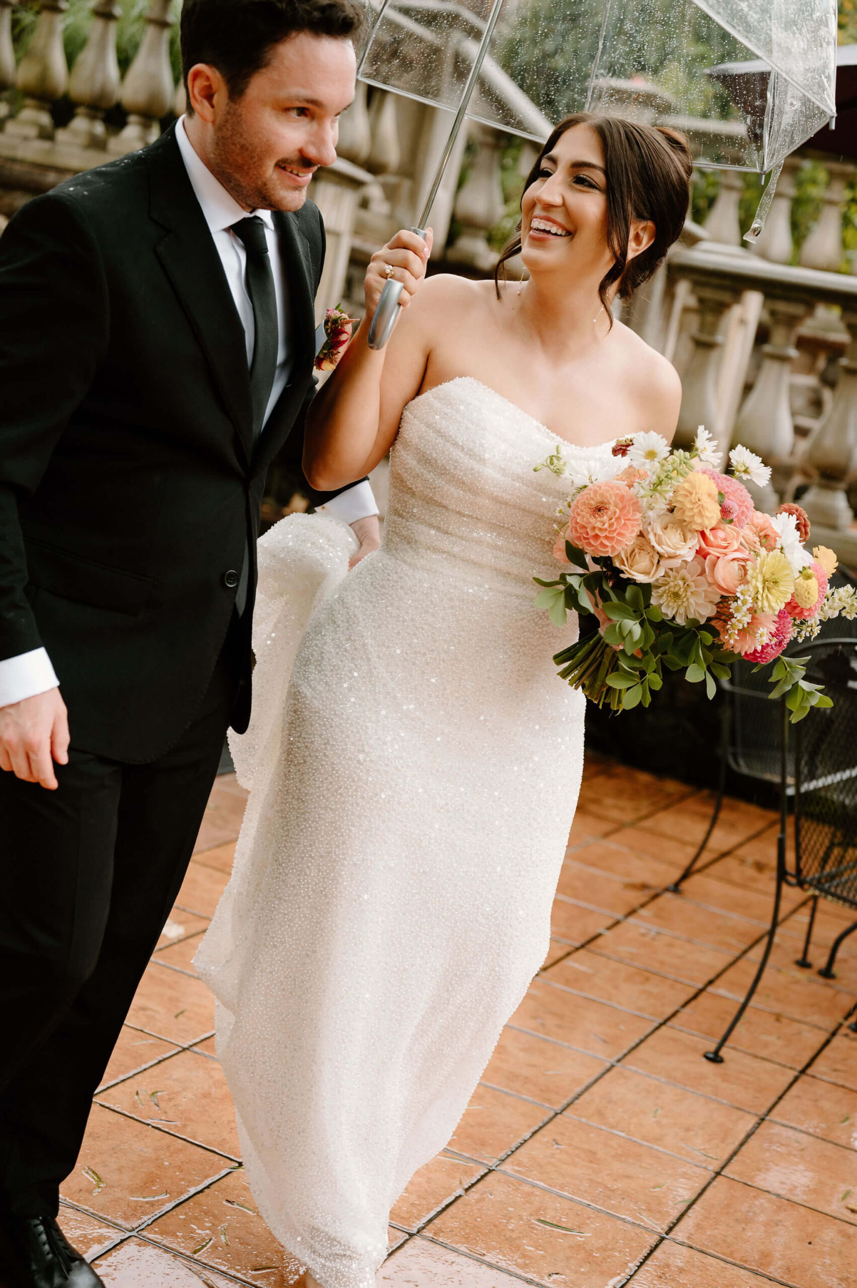 bride smiling at groom as they walk through rainy courtyard with an umbrella, groom holding the train of her sequins wedding gown