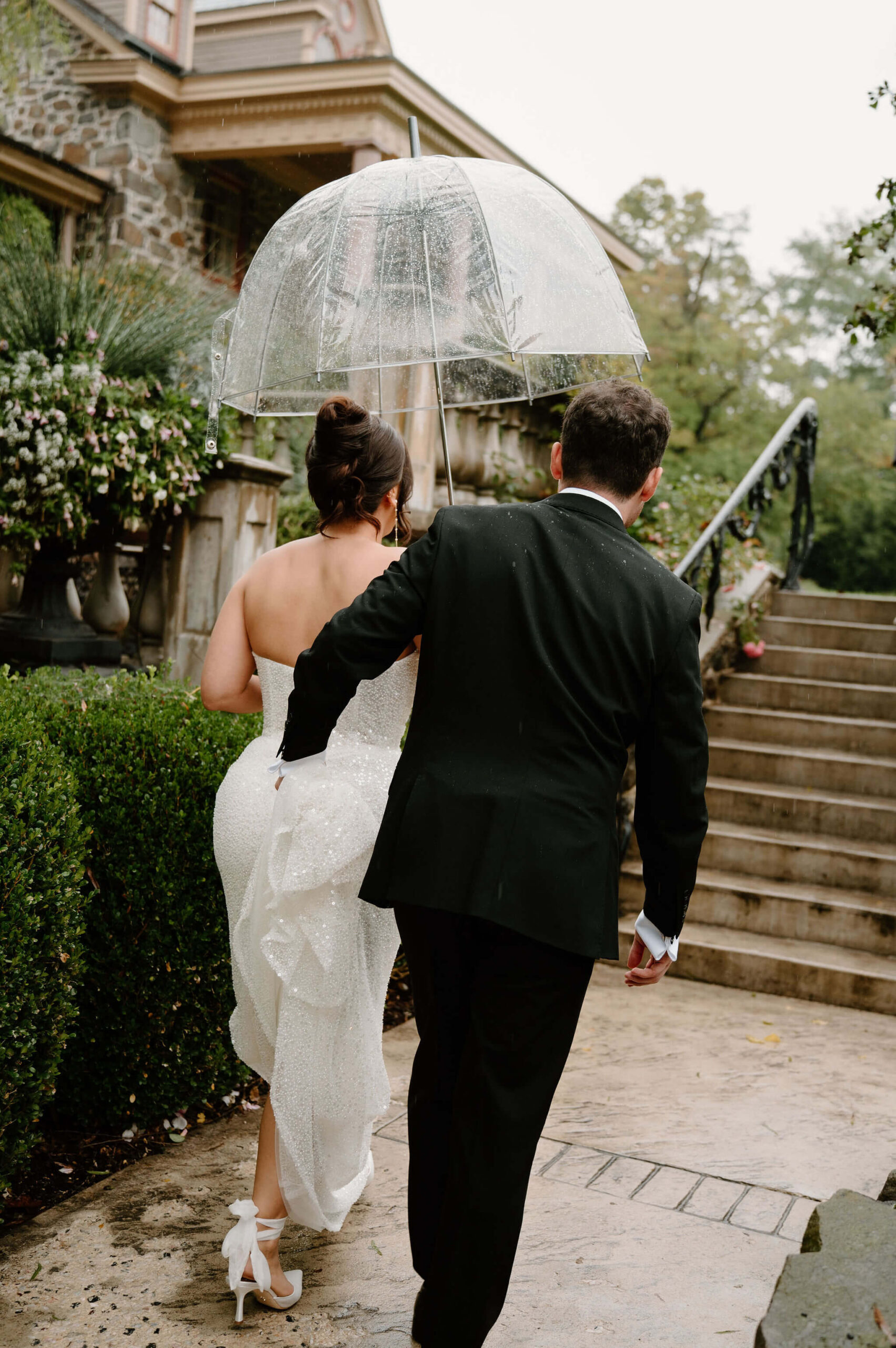 bride and groom walking along a path in the rain under a clear umbrella, groom holding the train of her sequins wedding gown