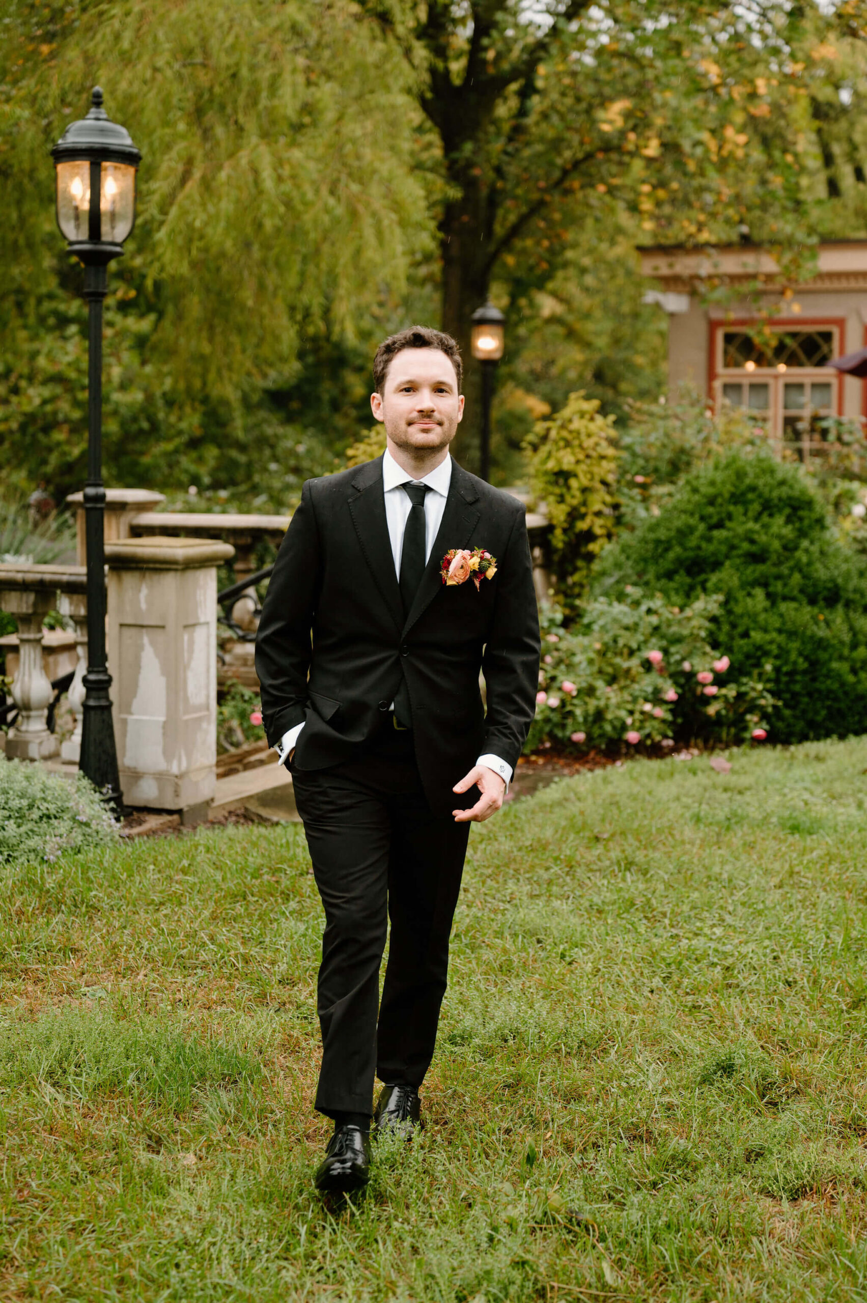 portrait of groom in a black suit with a colorful boutonniere, one hand in his pocket as he walks toward the camera with a soft smile on the grass
