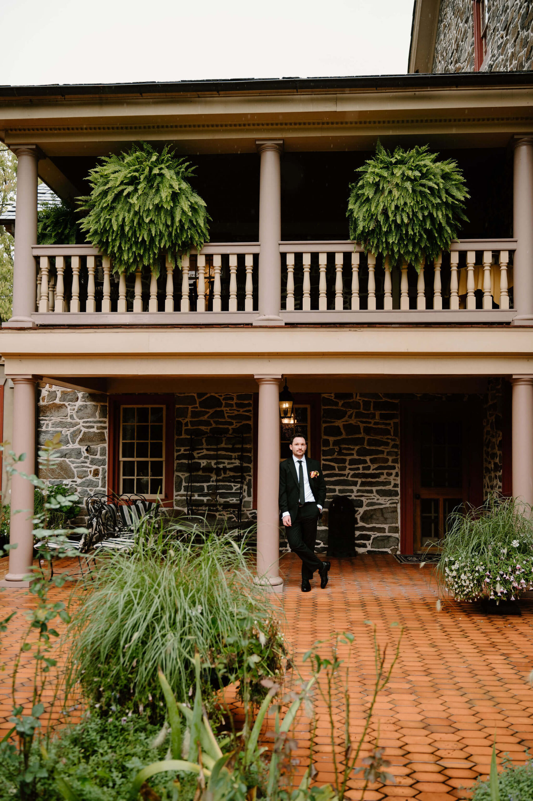 wide portrait of groom in a black suit leaning against a pillar on a covered porch, one hand in his pocket