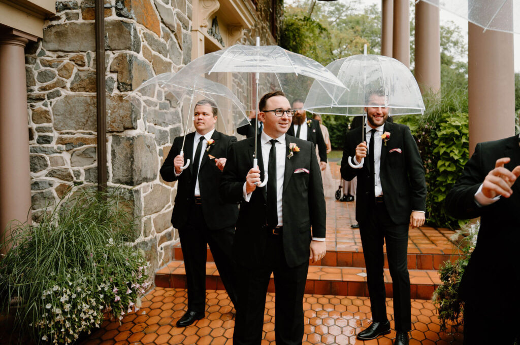 groomsmen in black suits carrying clear umbrellas walking along tiled patio