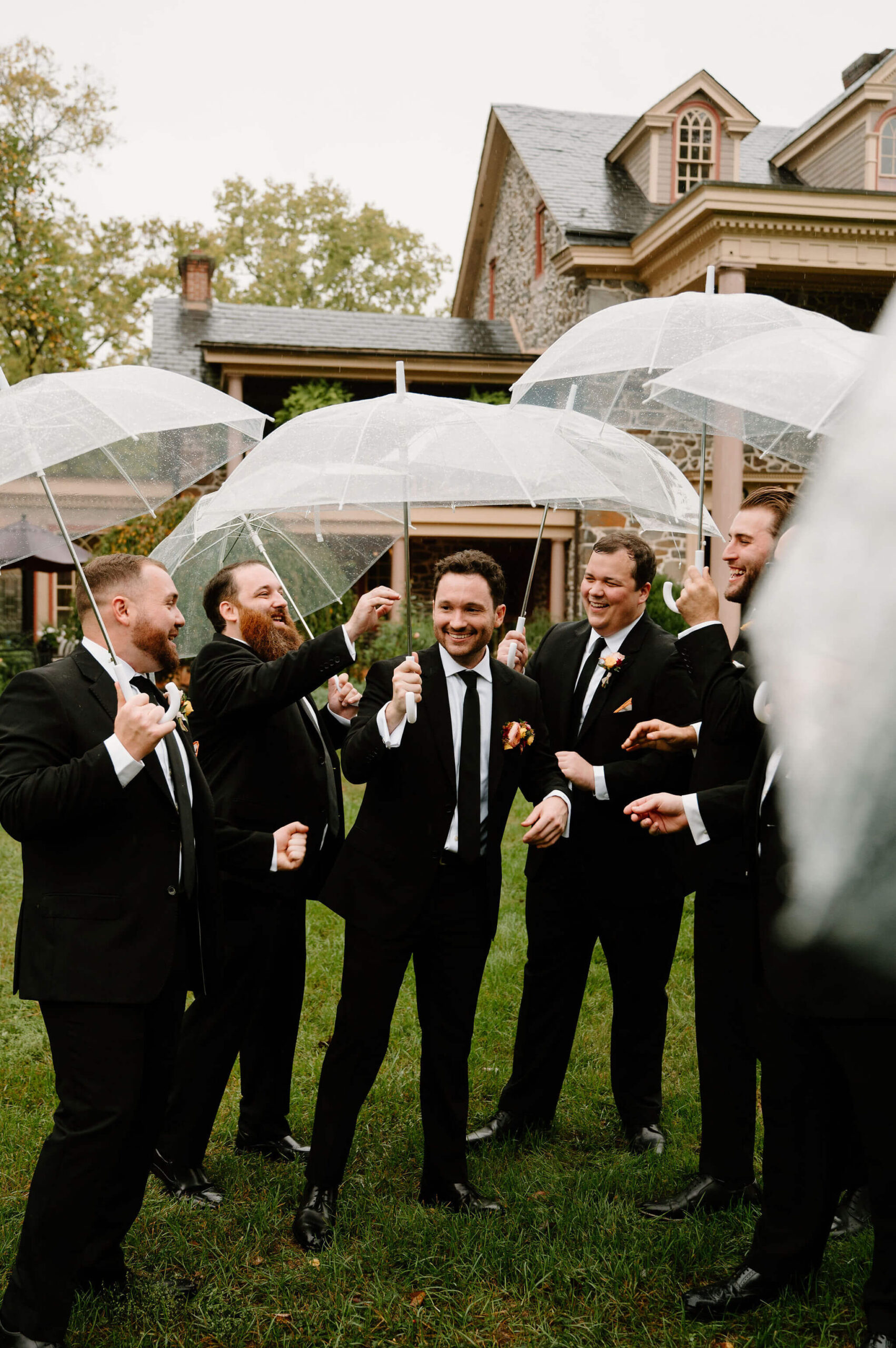 groom surrounded by groomsmen standing in a grassy yard, beneath clear umbrellas on a rainy afternoon, all smiling and talking together