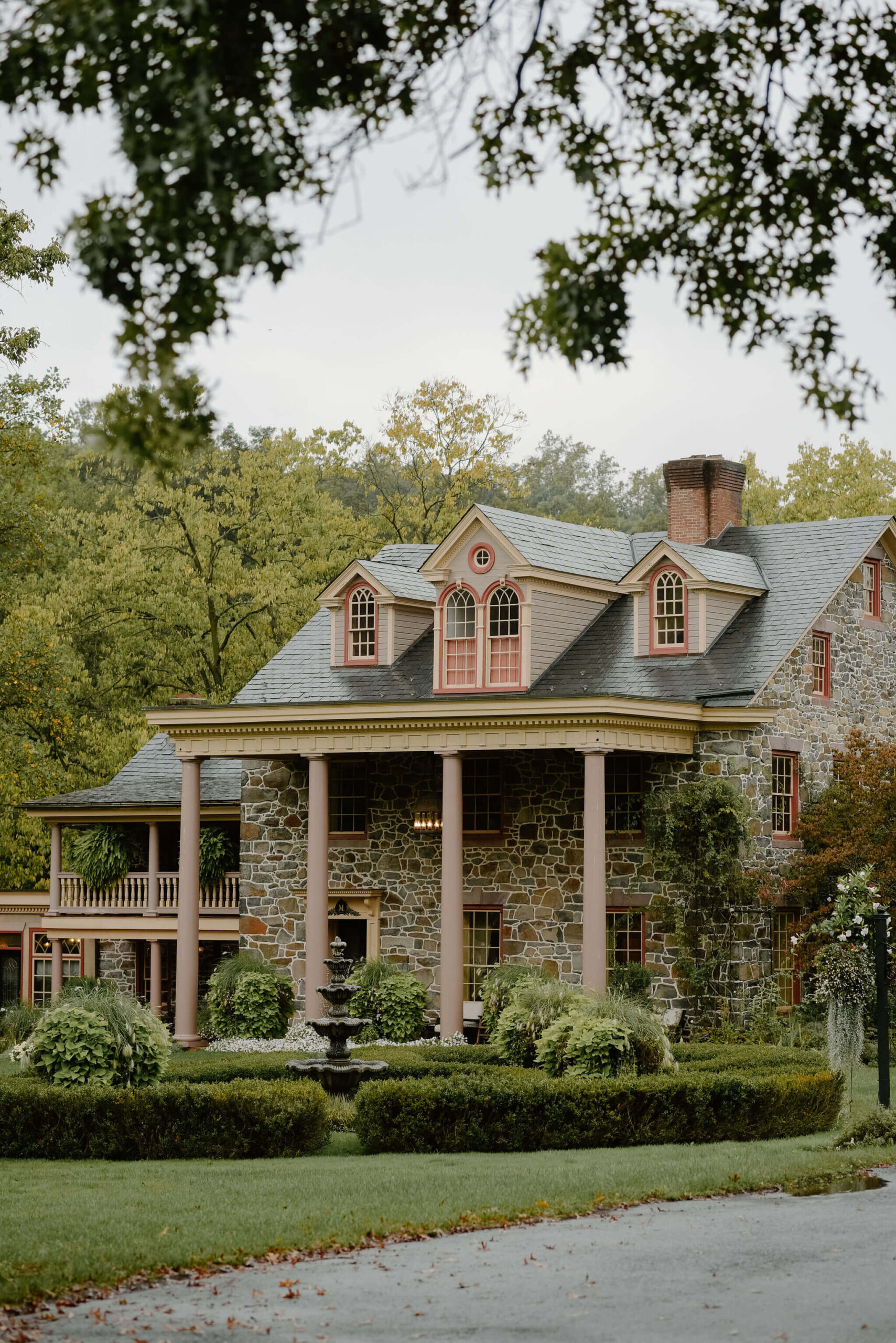 image of Moonstone Manor on a rainy September wedding day, a large stone estate with pillars holding up the ceiling of a porch, in front of a beautiful hedge row and fountain