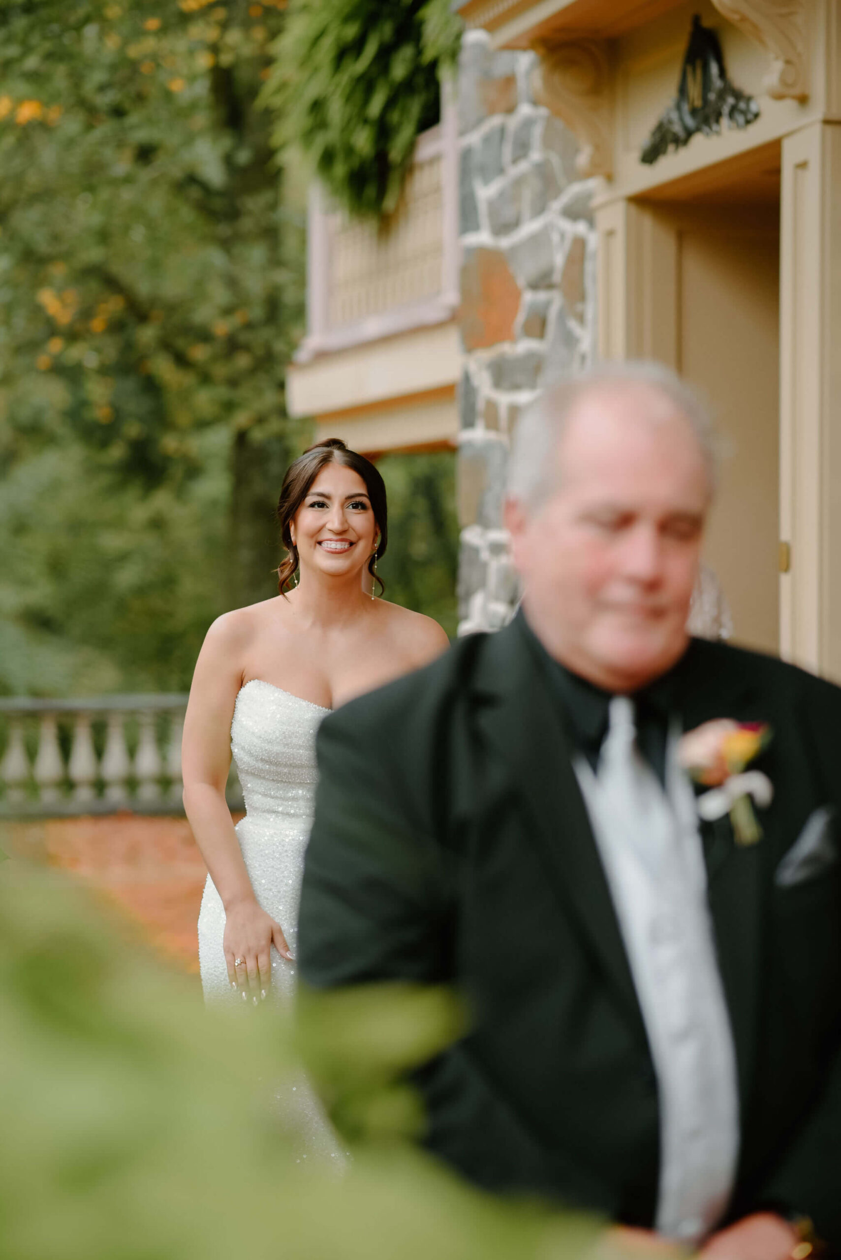 bride approaching her father (black suit, colorful boutonniere) for their first look, against a stone background