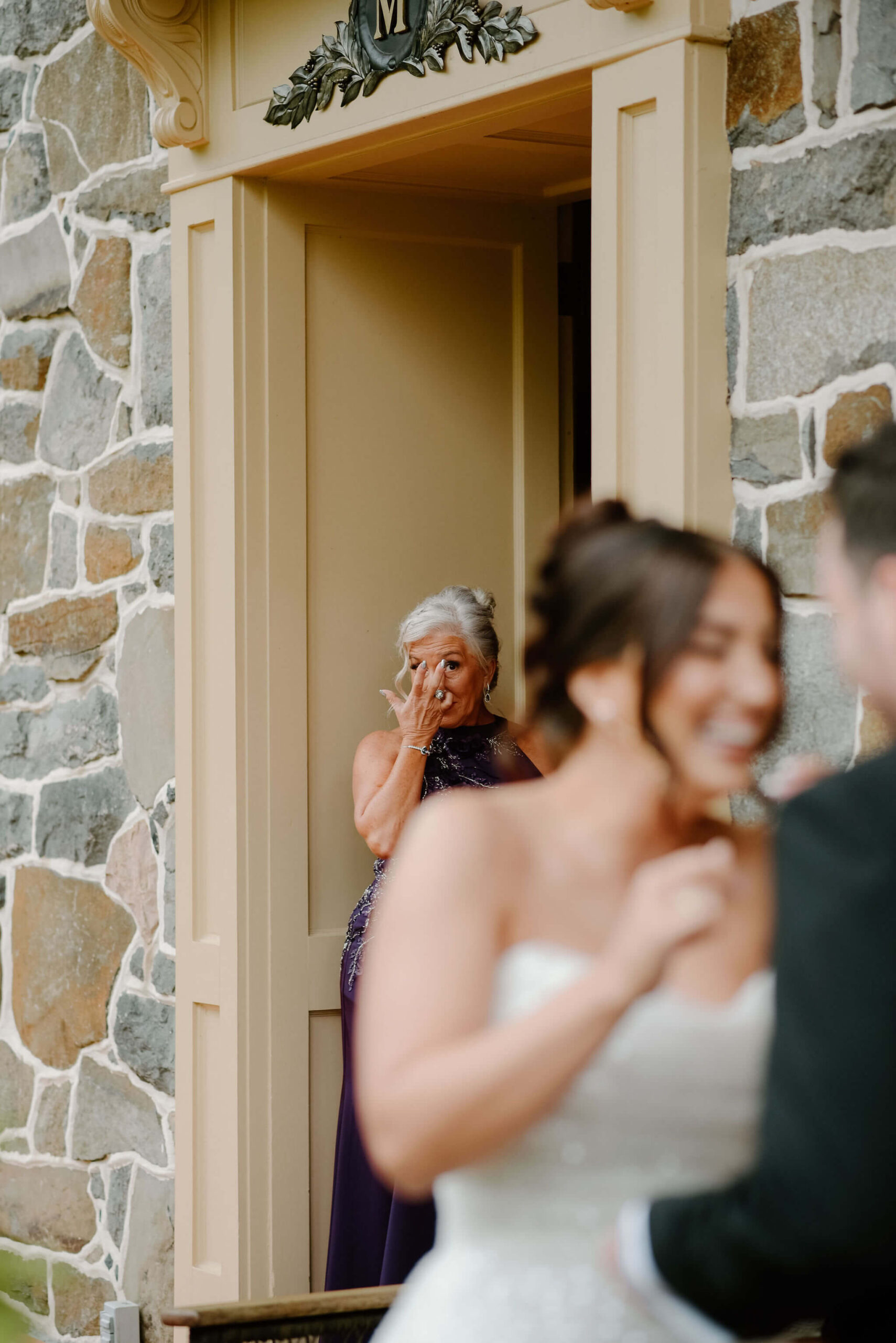 mother of the groom wiping her tears as she watches bride and groom's first look on their wedding day