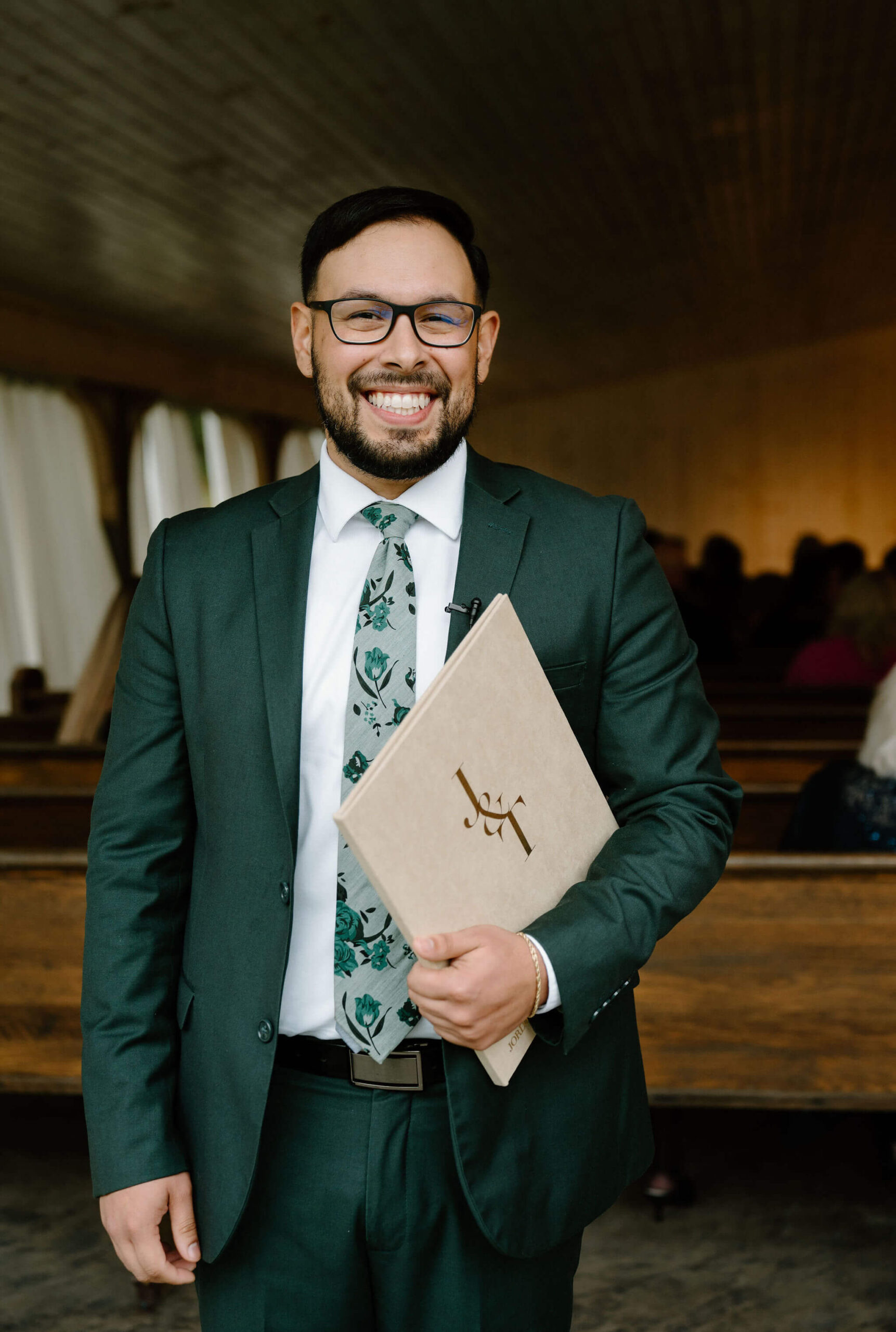 officiant in a green suit and blue-green floral tie, holding a binder labeled "J&T" and smiling at the camera