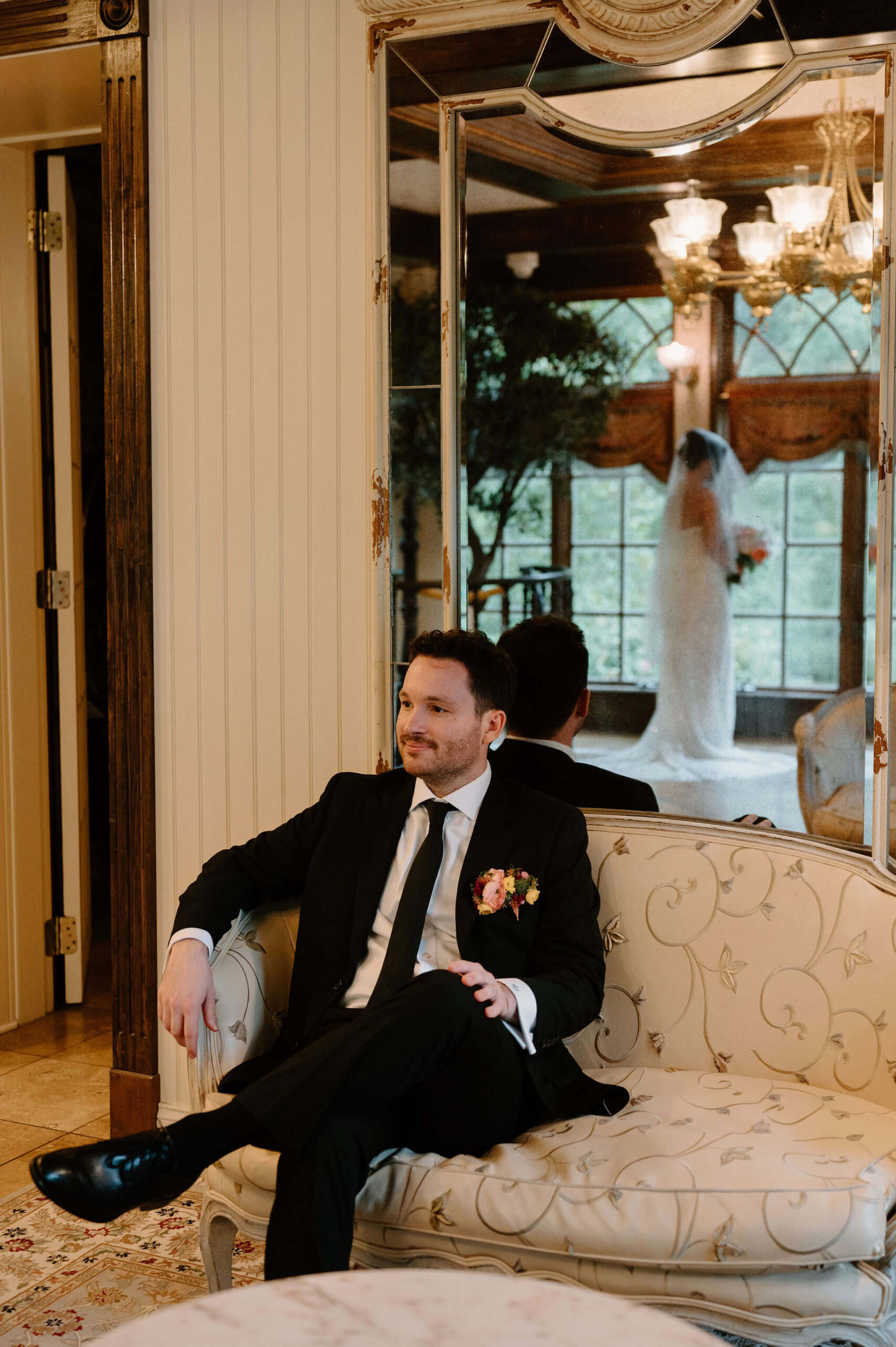 groom (black suit, colorful boutonniere) seated on an embroidered couch and smiling softly on camera. behind him, reflected in a mirror, the viewer can see his bride in a sparkly white gown and veil, posing for solo portraits with her flowers