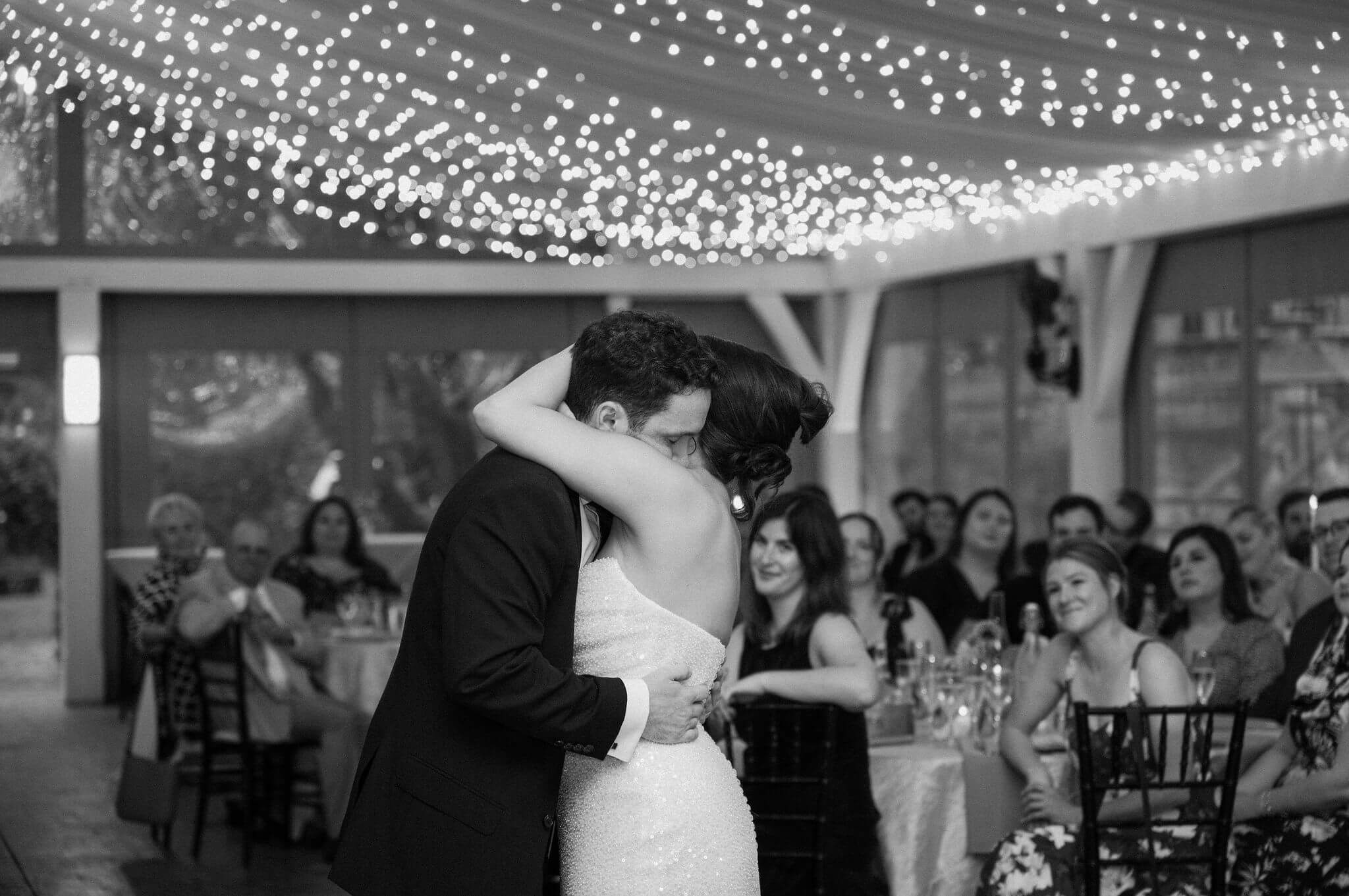 black and white image of bride and groom embracing, eyes closed, during their first dance while guests watch joyfully