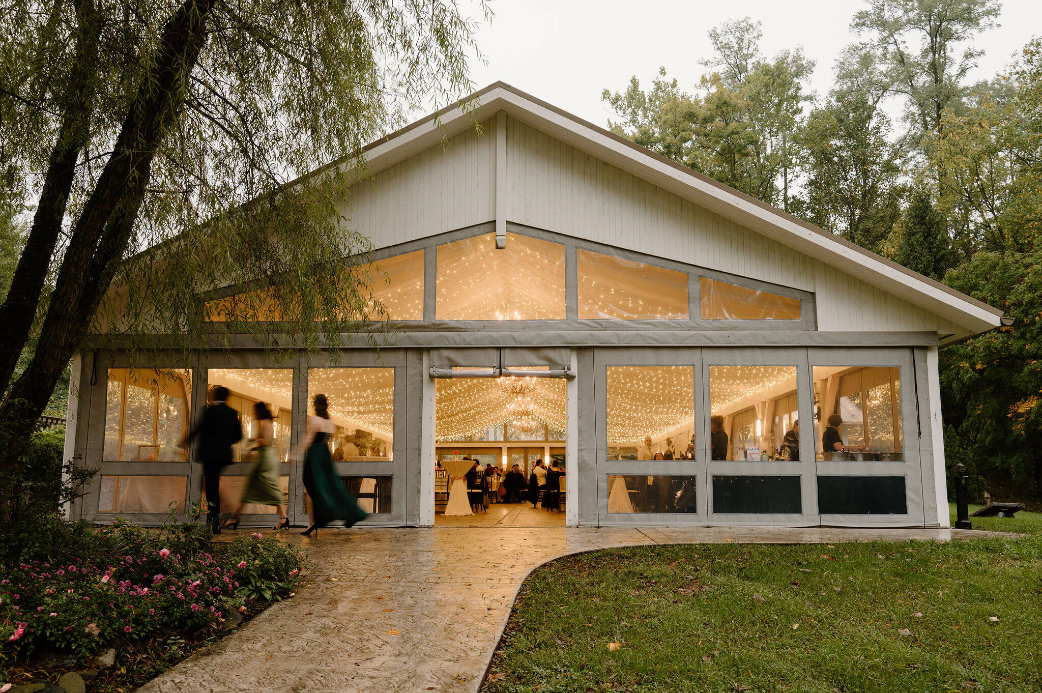 wide image of a permanent tent structure with clear walls and lots of string lights on a rainy wedding day, guests walking in front of the structure