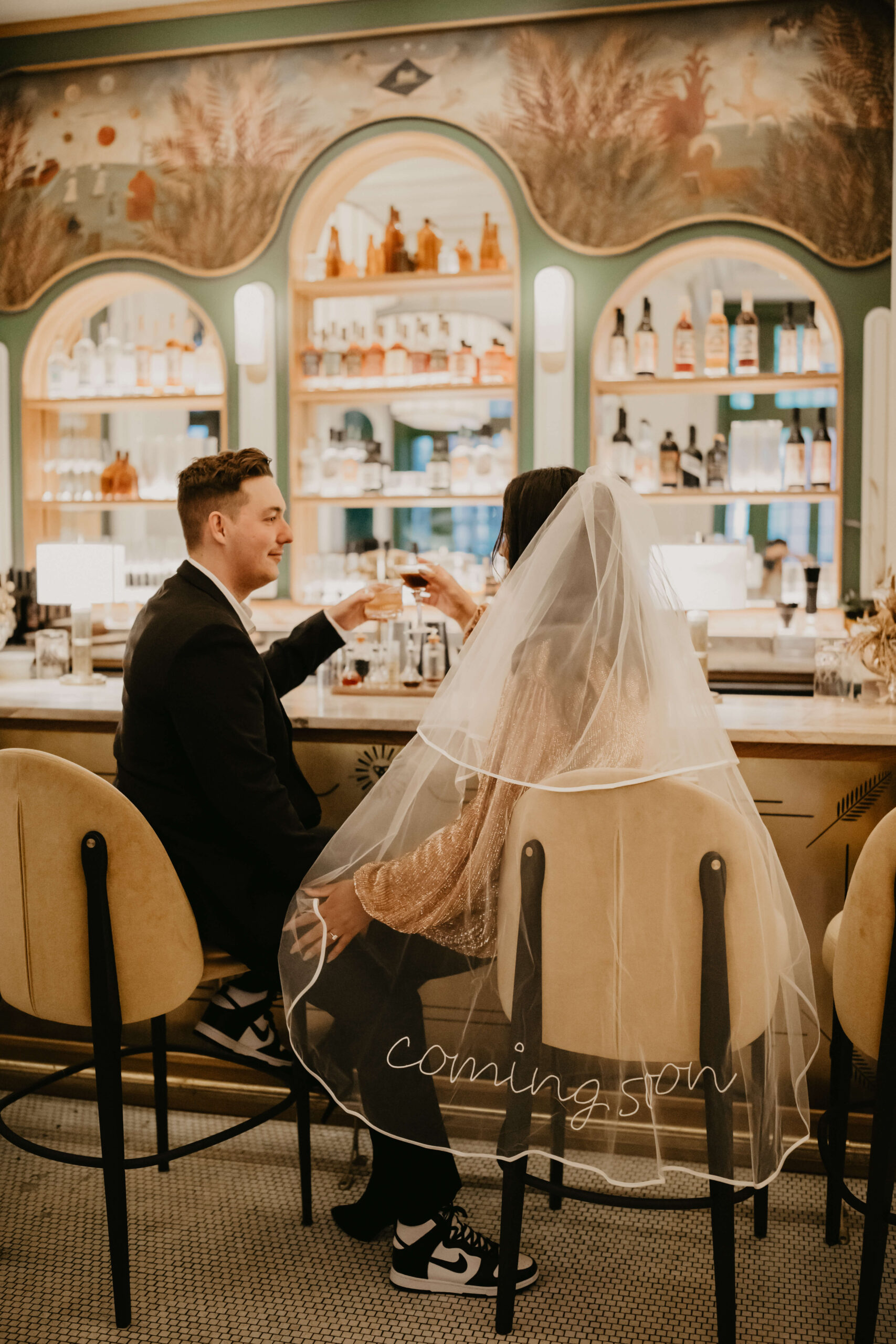 engaged couple sitting at a beautiful cocktail bar, clinking cocktail glasses, with the bride wearing a veil embroidered with the words "coming soon"