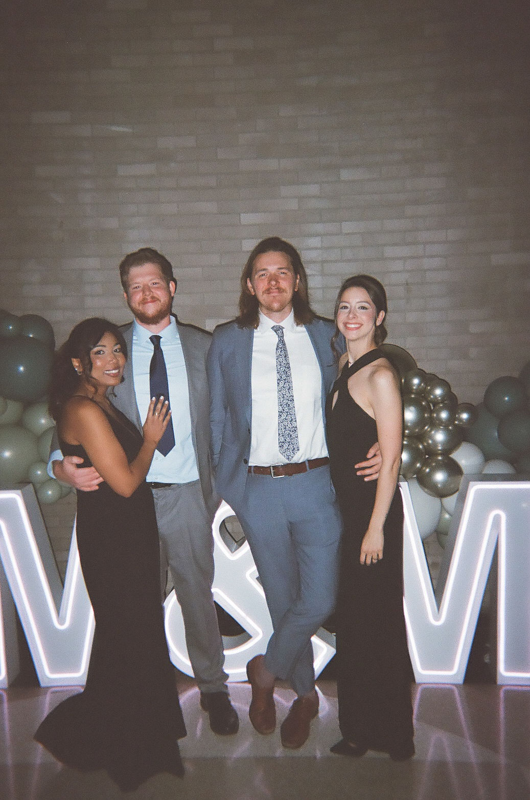 two couples wearing formalwear smiling at the camera during cocktail hour, in front of a large lit-up sign that says "M&M"