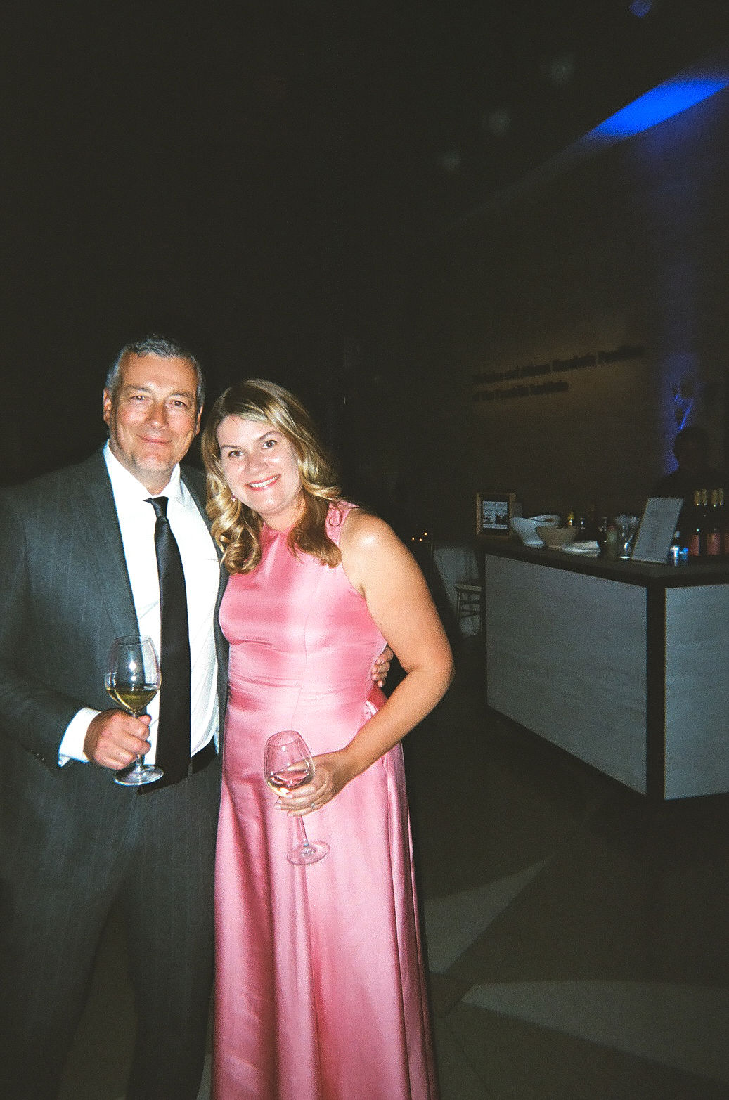 a couple smiling at the camera while holding wine glasses, wearing formalwear during a wedding cocktail hour
