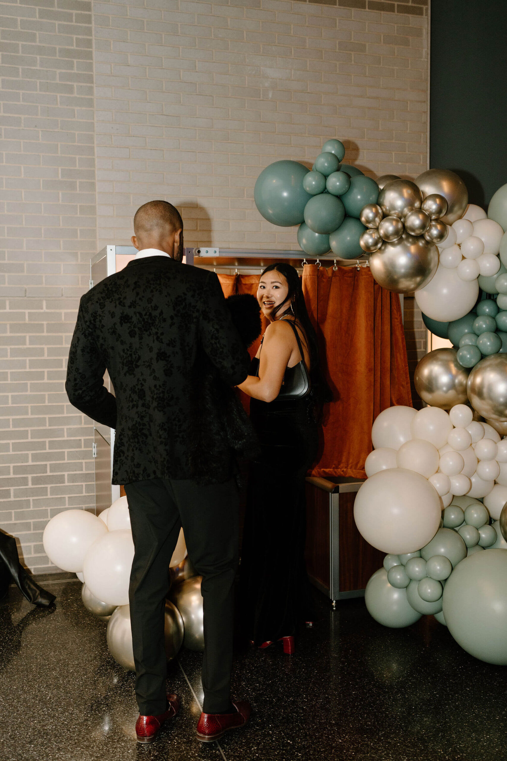 two guests in formalwear stepping into a photobooth, surrounded by a large white and green balloon installation