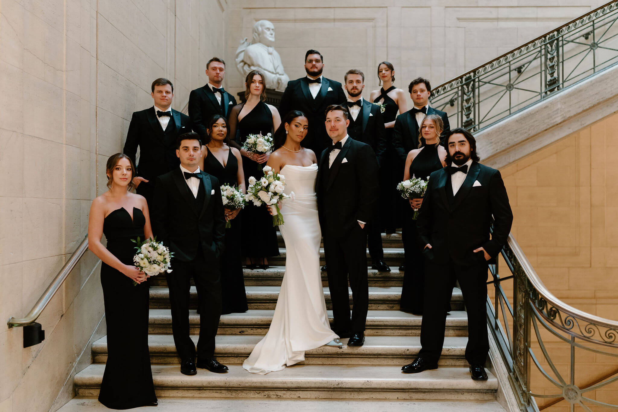 stoic image of bride and groom with their full wedding party at their franklin institute wedding, at staggered heights on a marble staircase, all wearing black except the bride and looking right at the camera