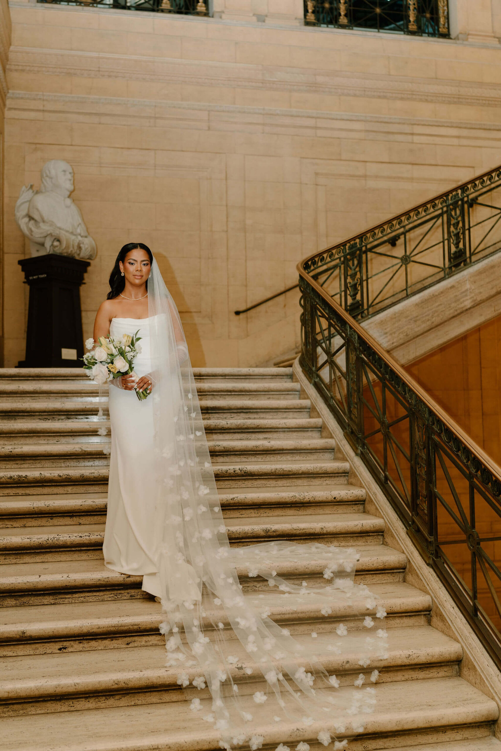 bride standing on a very large marble staircase, holding her wedding bouquet in her hands and smiling softly at the camera, with a cathedral length veil flowing down the stairs