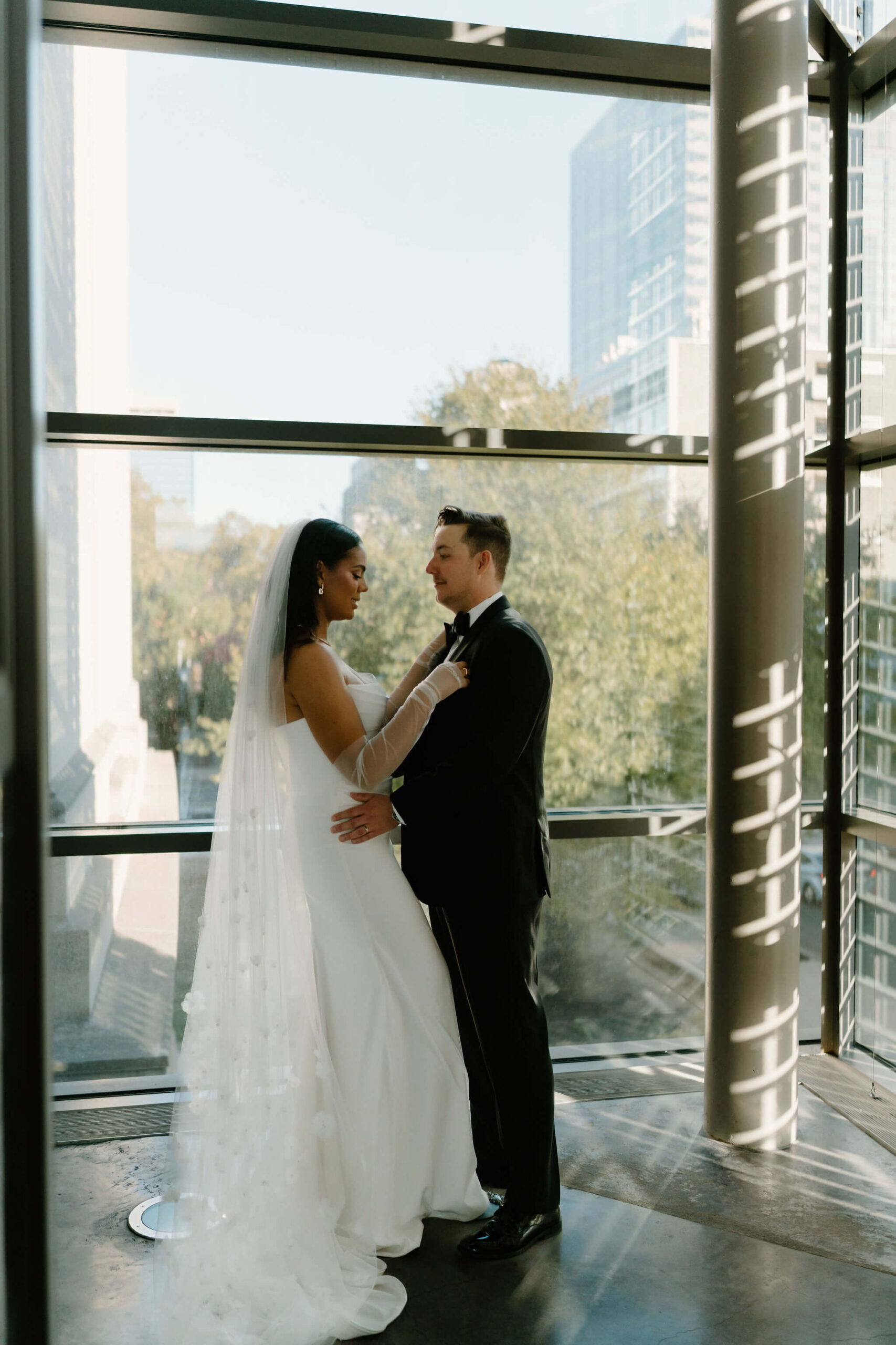 bride and groom in front of a large window, embracing, while she holds his lapel at their franklin institute wedding