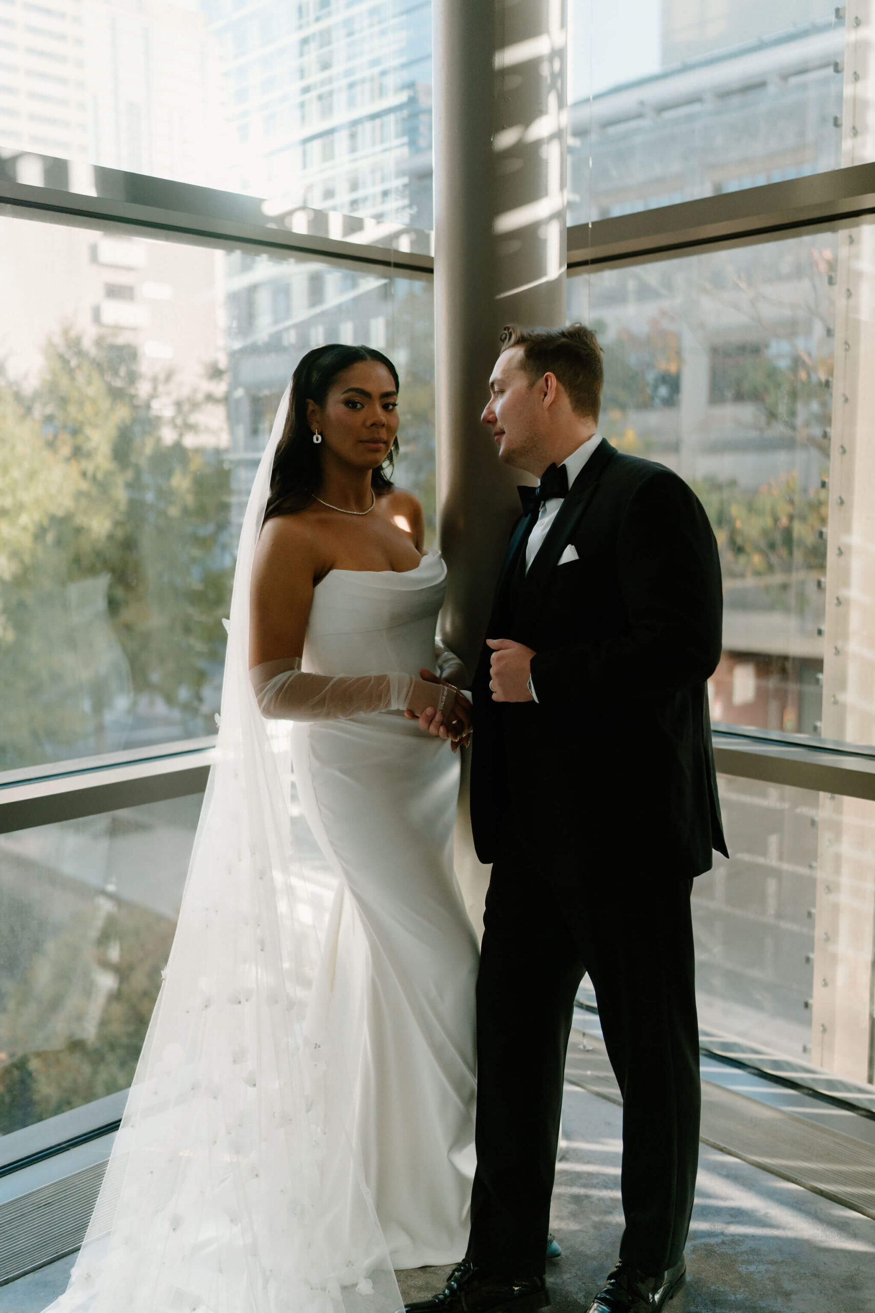 bride and groom embracing in front of a large window, bride looking at the camera