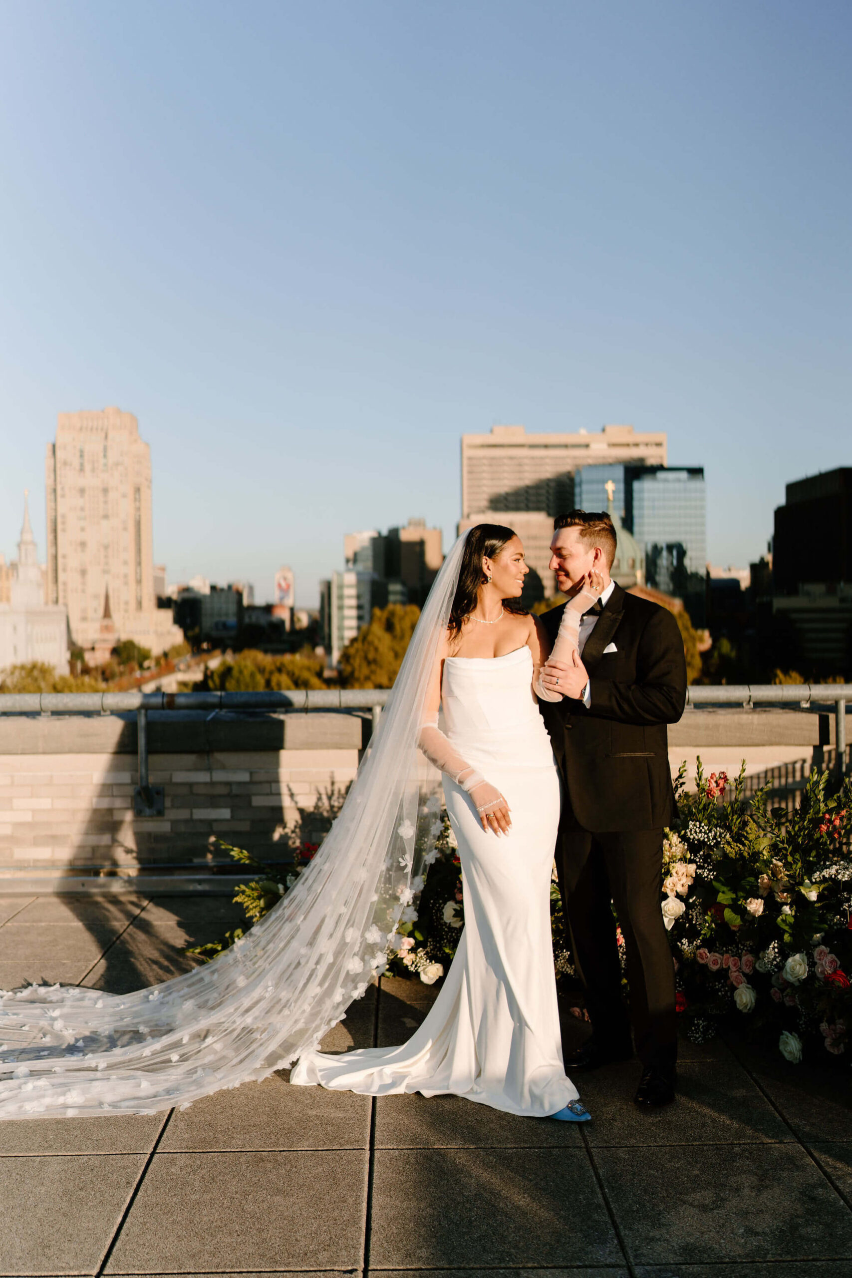 wider image of bride and groom looking at each other and smiling while standing side by side on the roof of the Franklin Institute, her left hand reaching up to hold his face and his left hand on her forearm